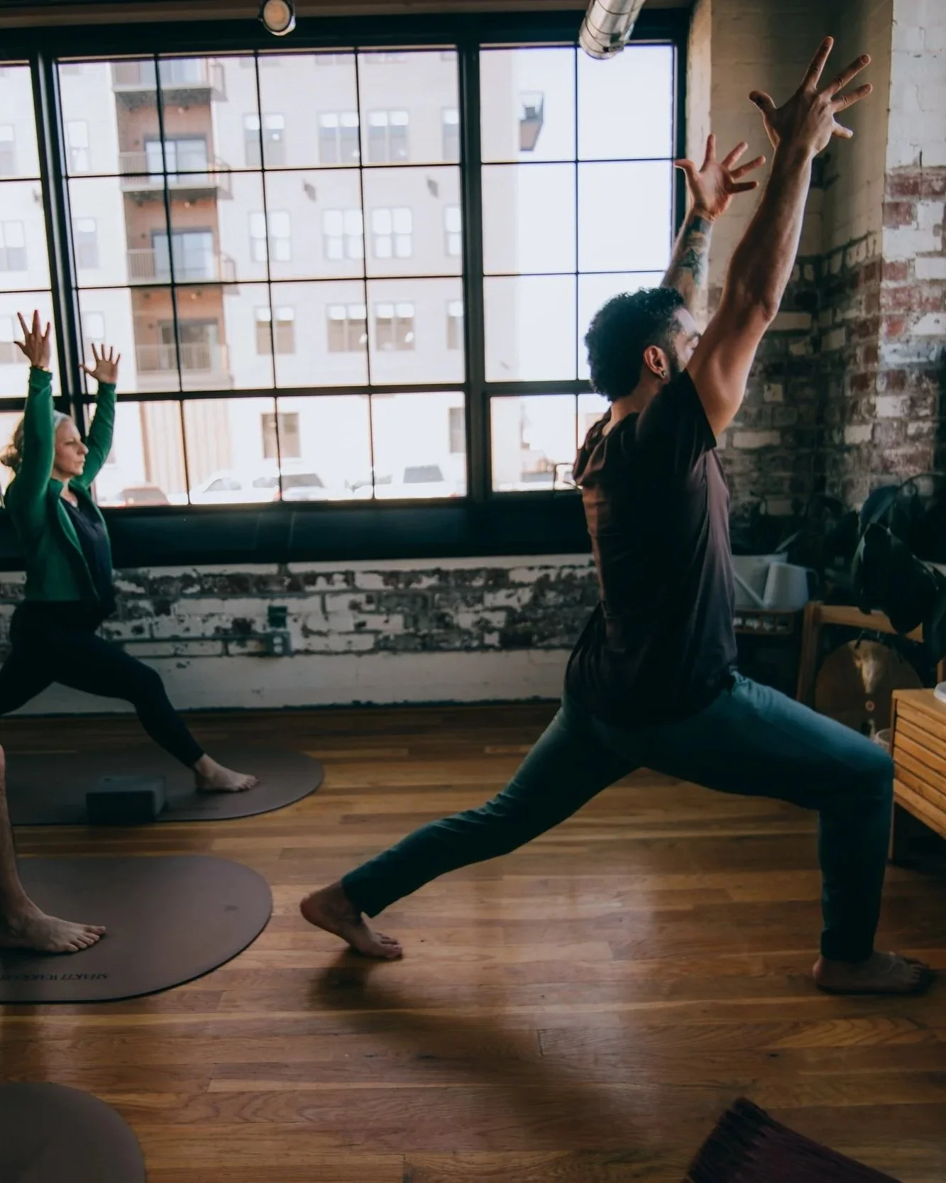 Two people practicing yoga in a spacious studio with large windows and exposed brick wall, both in Warrior pose with arms raised and legs apart.