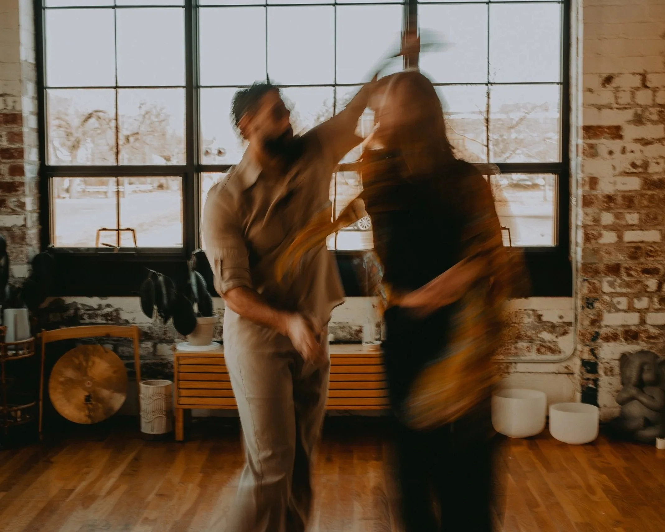 Two people dancing passionately in a studio with large windows and brick walls. The image is blurry due to motion.