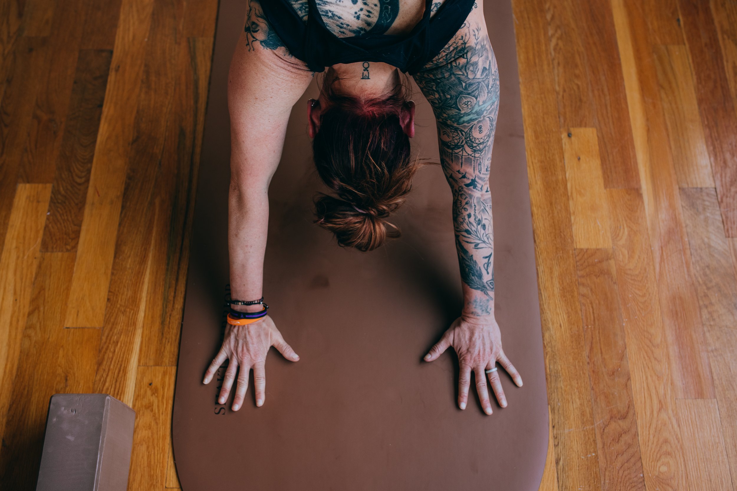 A woman with tattoos on her arms and neck practicing yoga in downward dog pose on a brown mat on a wooden floor.