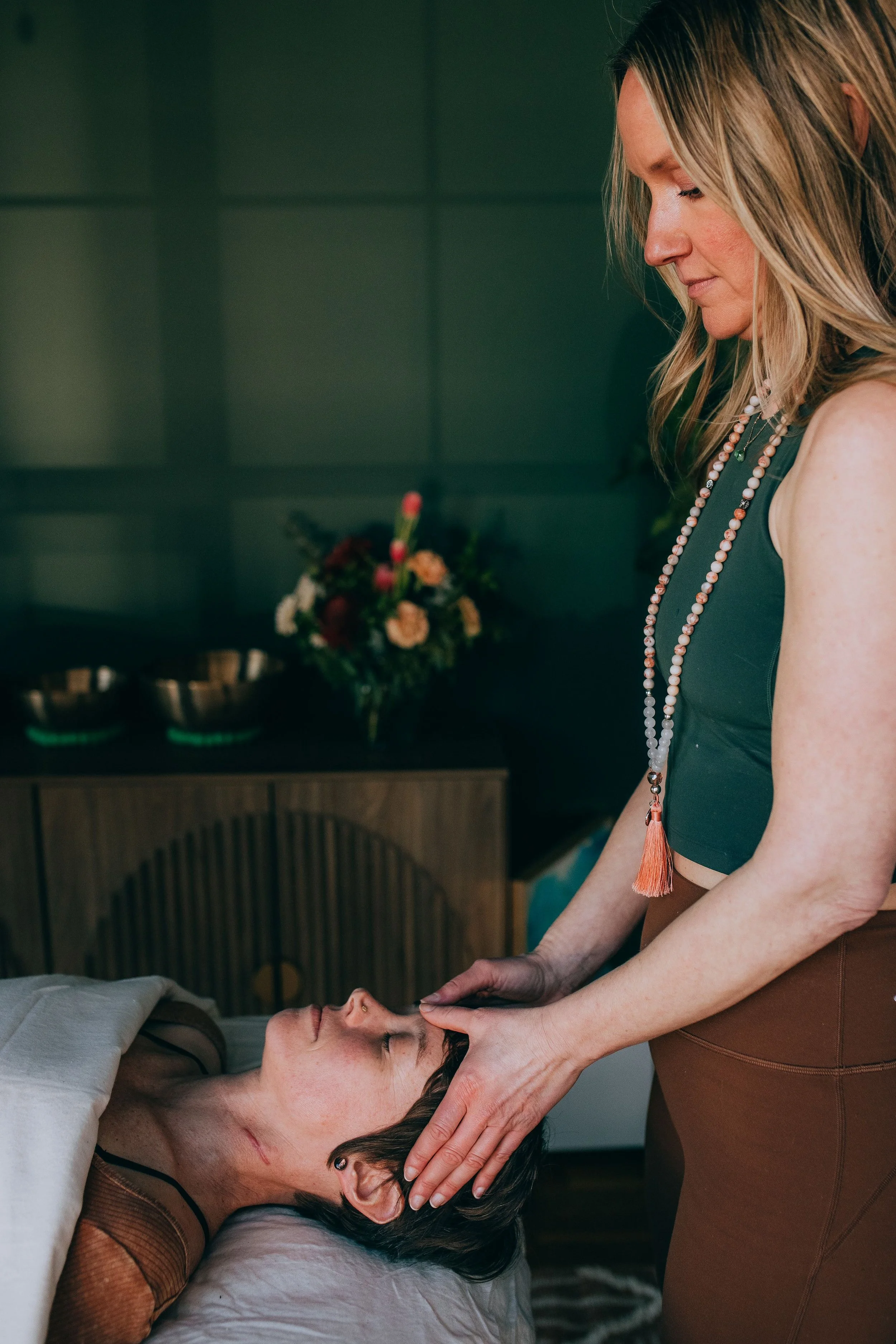 A woman with long hair wearing a beaded necklace and sleeveless top is giving a massage to a person lying down, indoors with a dark green wall and a bouquet of flowers in the background.