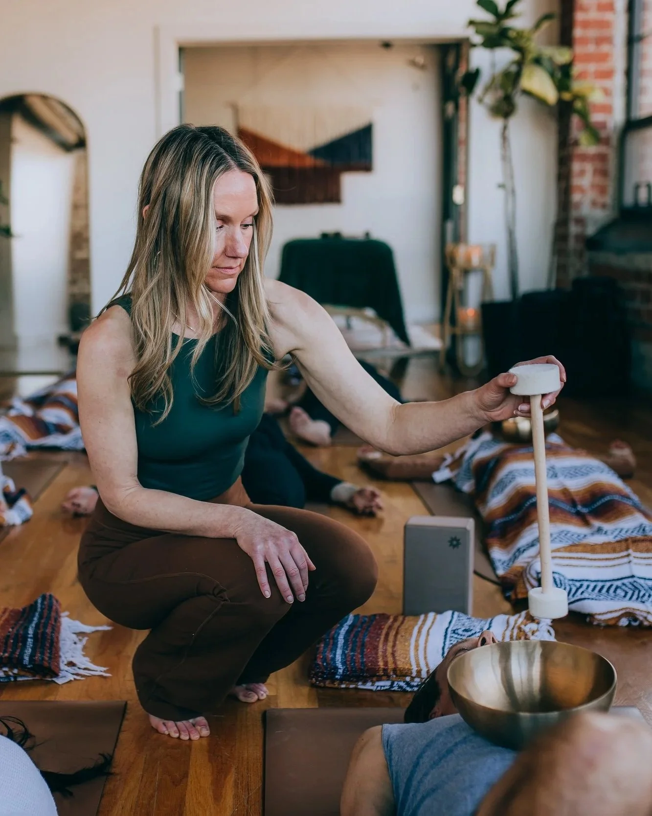 A woman practicing sound healing or meditation using a crystal singing bowl in a cozy indoor setting with other participants lying on the floor on yoga mats and colorful blankets.