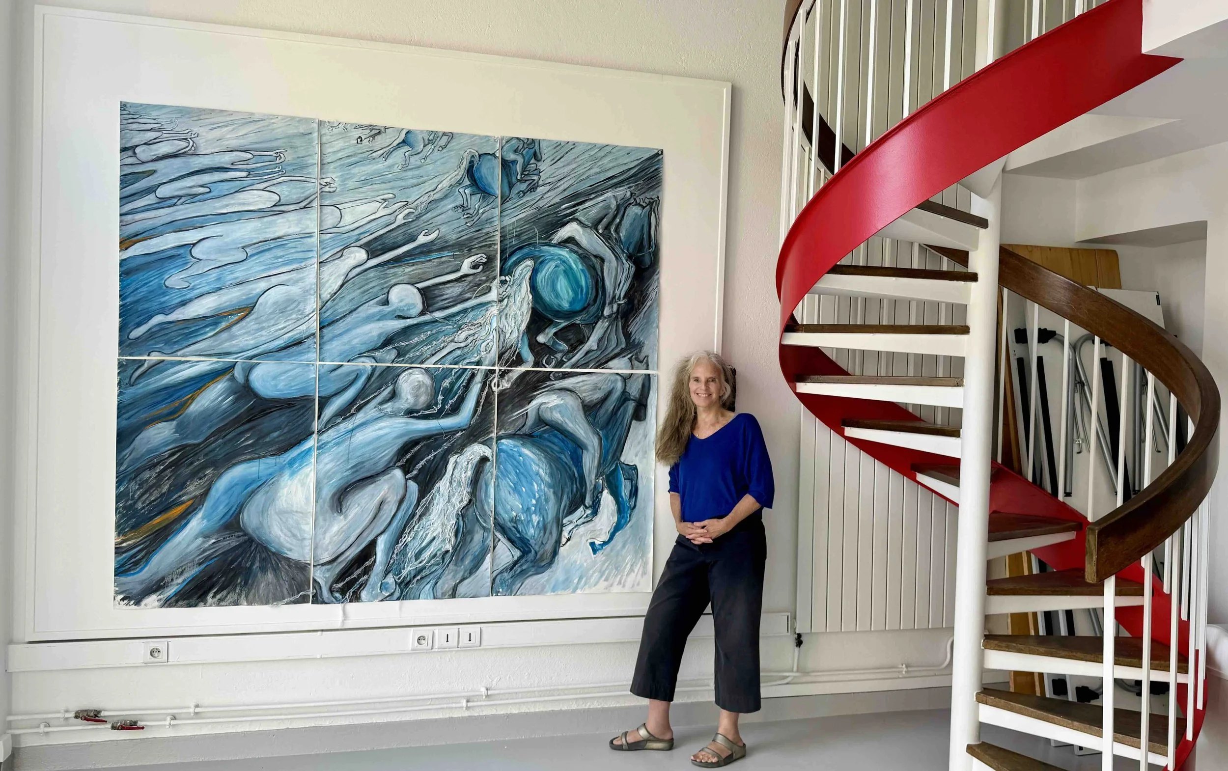 A woman with long white hair in a blue blouse and black pants standing next to a large abstract painting with blue and white colors. She is indoors in front of a red spiral staircase and white wall.