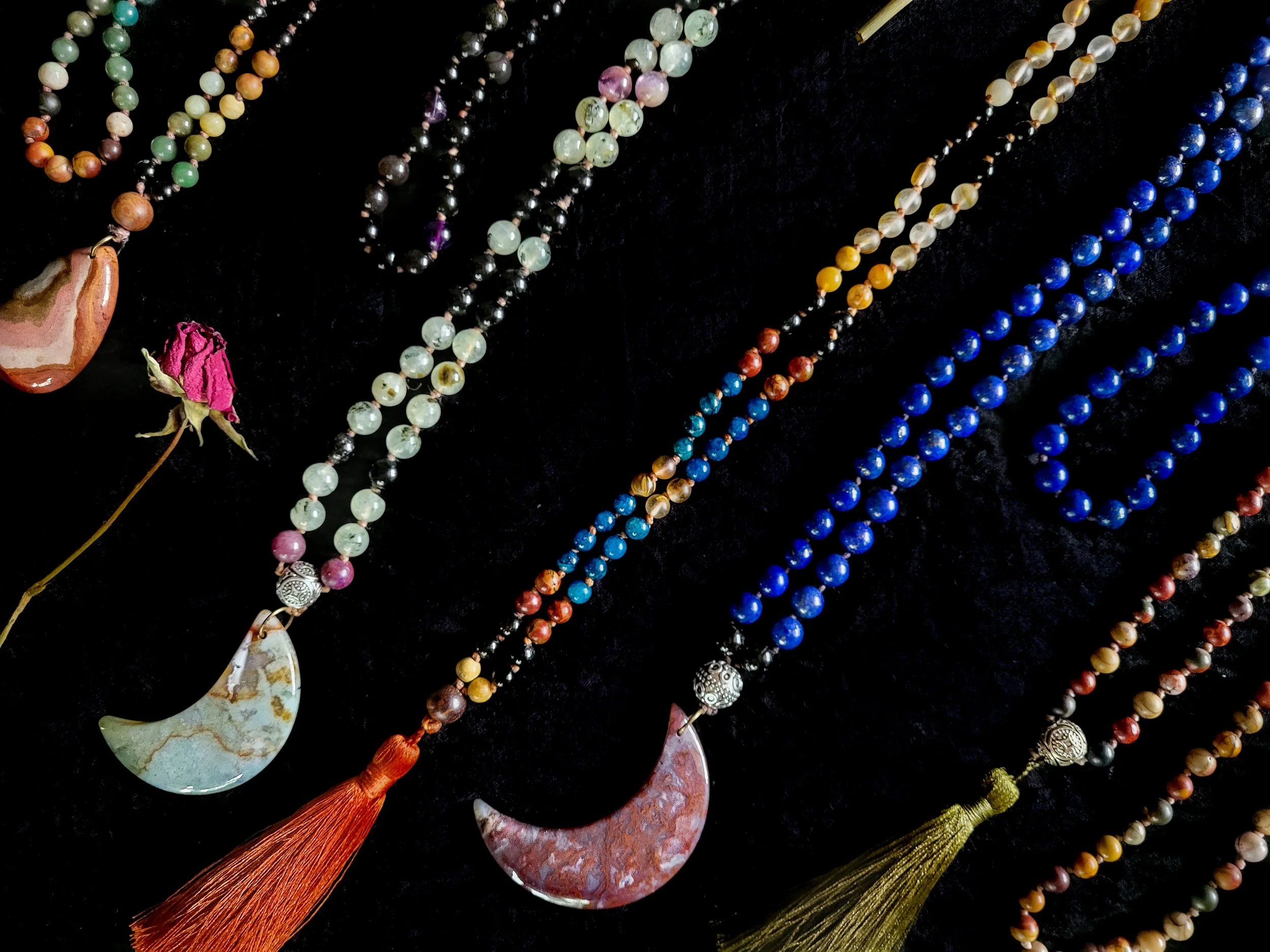 Photo of four custom-made limited gemstone mala necklaces with 8mm and 6mm beads; polychrome jasper and agate gemstone pendants; and a dark orange and an olive green silk tassel. Beads include dark blue lapis lazuli, green and yellow jade, amazonite,