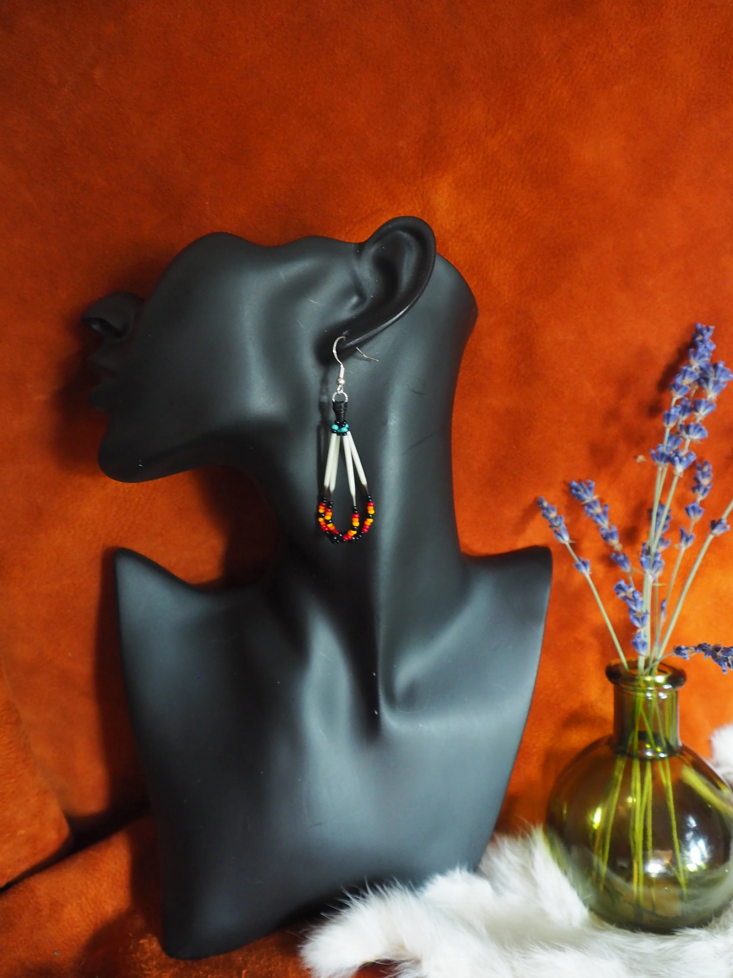 Photo of small Wabanaki Porcupine Quill earring hung on a dark jewelry mannequin in front of dark terra cotta colored leather backdrop, next to a green round glass jar with lavender and white fur.  Earrings contain four strands of two looped segments