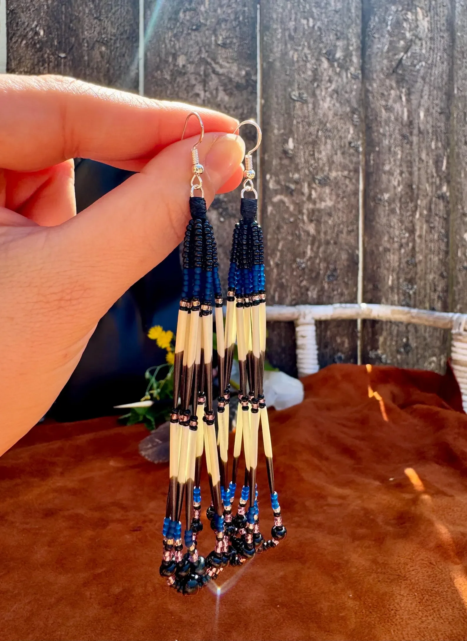 Photo of two long, lightweight Porcupine Quill earrings hung dangling from the artist's fingers in front of a weathered light brown wooden fence, with a white table covered in dark terra cotta colored leather. Each earring is comprised of 8 strands o