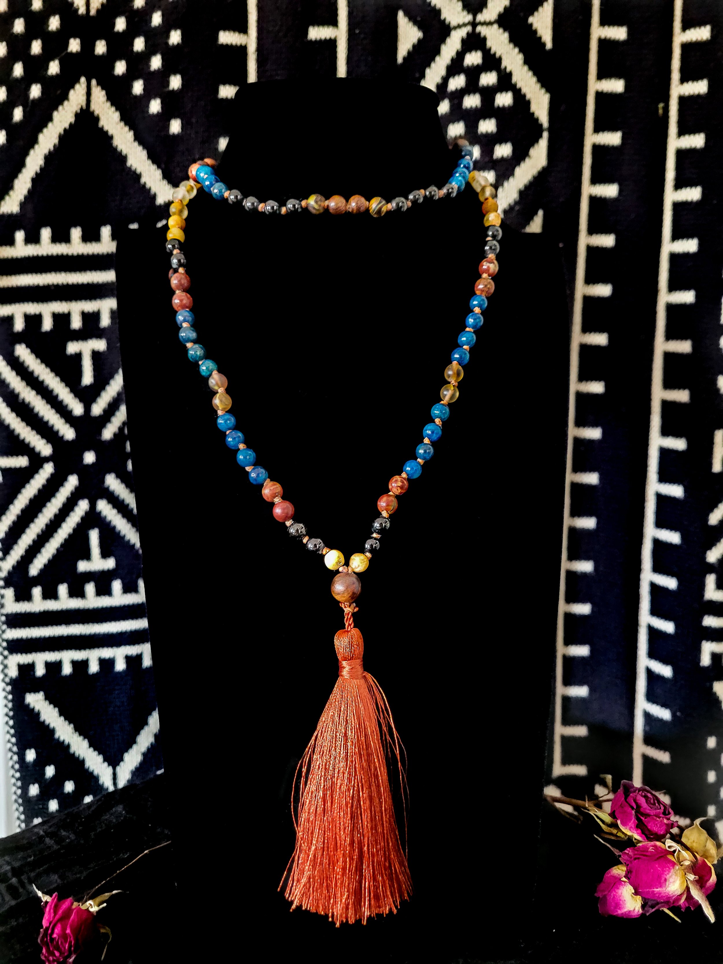 Photo of custom-made limited gemstone mala necklace with 8mm tourmaline, yellow jade, amazonite, picture and crazy horse jasper, fire agate, lapis lazuli; 6mm sandalwood bead; and dark orange silk tassel. Mala is shown on a dark necklace display with