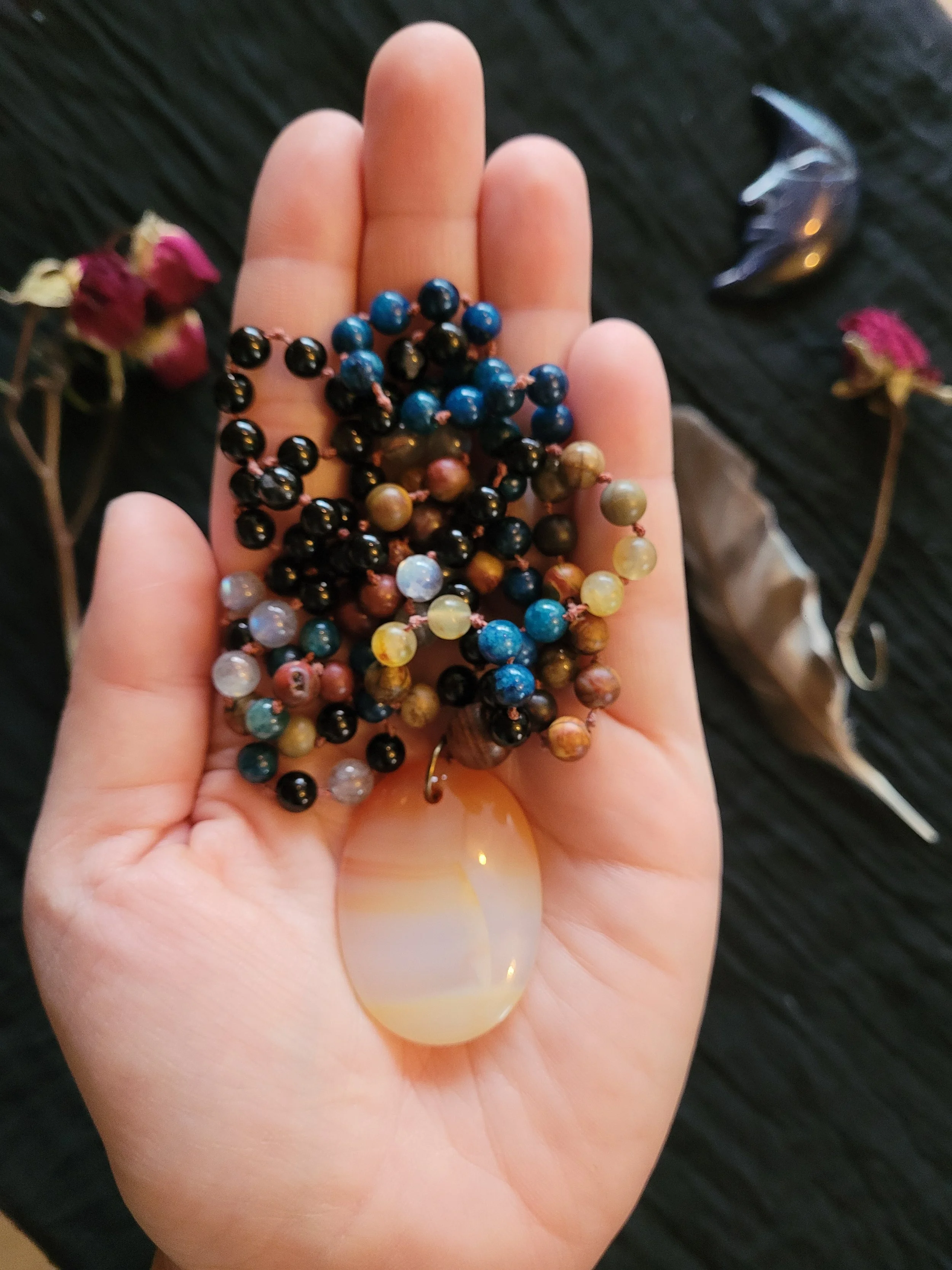 Photo of hand holding custom-made limited gemstone mala necklace with 8mm labradorite, lapis lazuli, crazy horse Jasper, tourmaline; 10mm sandalwood bead; and carnelian pendant. Feather, crystal, and dried roses are in the background. Commission desi