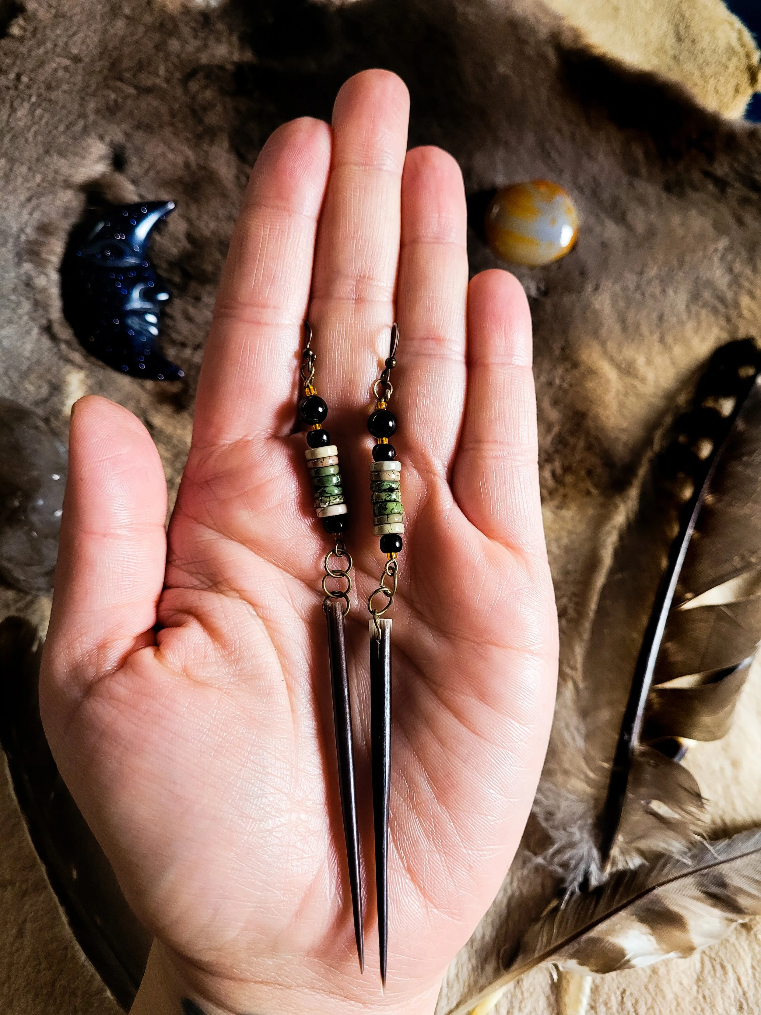 Photo of medium length, lightweight  African Porcupine Quill earrings held in the Artist's hand, with brown fur, crystals, and feathers in the background. Earrings contain two long  brown, sharply tipped African porcupine quills, picture Jasper beads