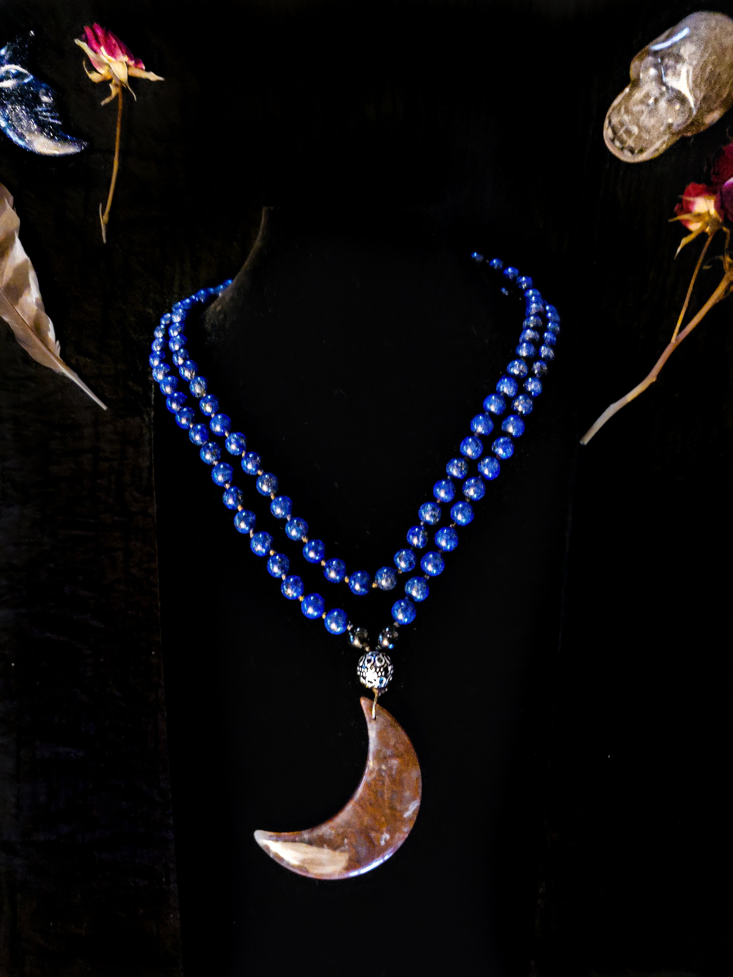 Photo of custom-made limited gemstone mala necklace with 6mm lapis lazuli; 4mm ornate silver finishing bead; and a large dark brown moss agate moon pendant. The mala is displayed on a dark black necklace display with a feather, crystal skull, blue cr
