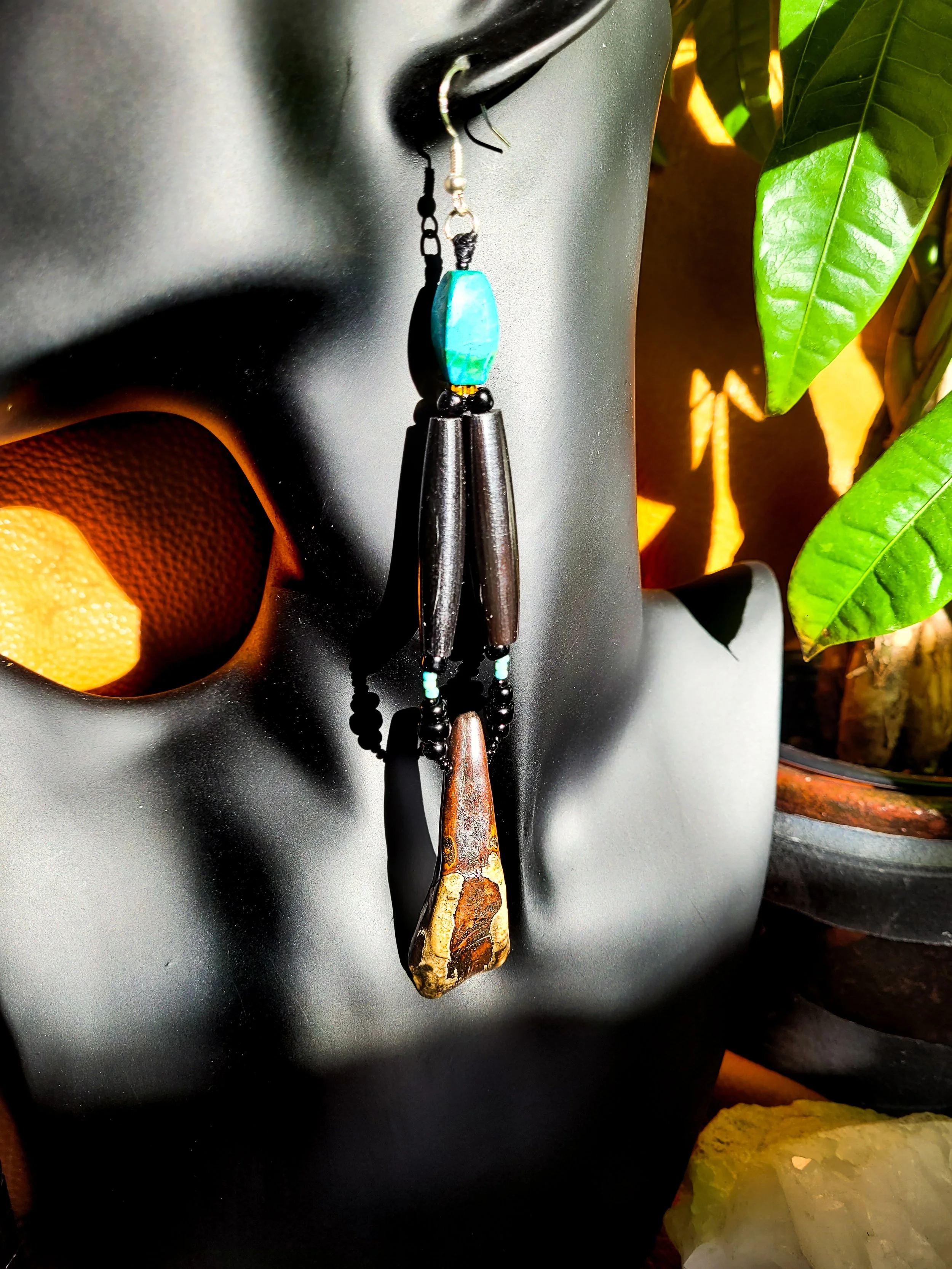 Photo of long, heavy-weight Turquoise, Bone, and Buffalo Tooth earrings displayed on a dark jewelry mannequin with tan leather, crystals, pottery, and a green plant in the background. Earrings contain two dark brown bone pipe beads, Buffalo tooth, re