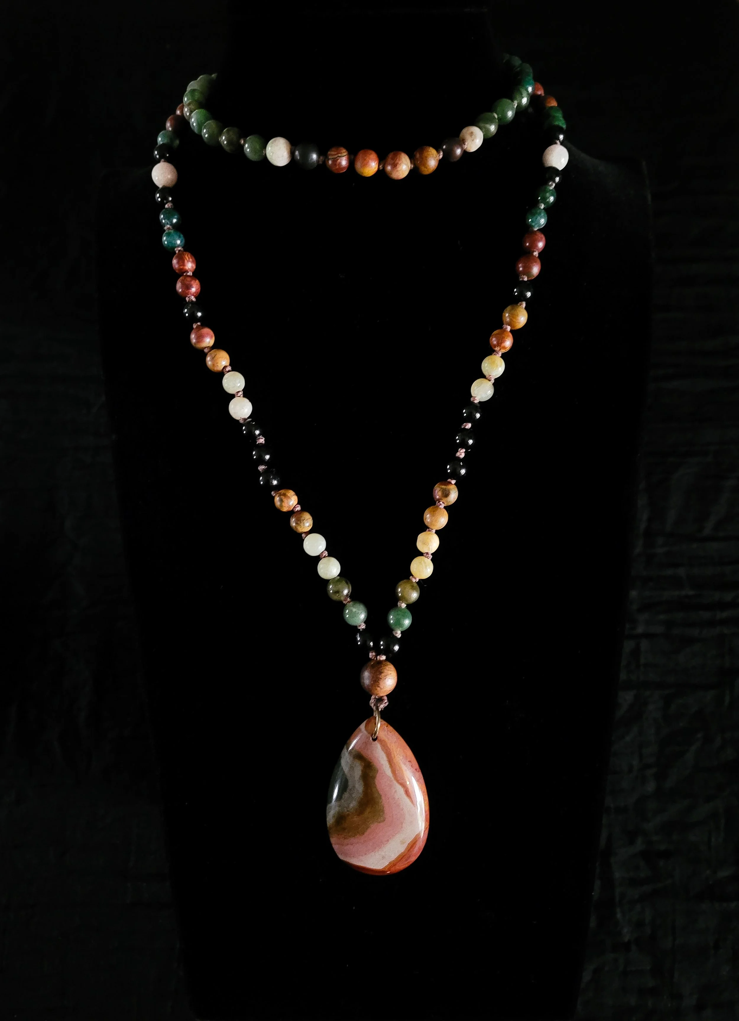 Photo of custom-made limited gemstone mala necklace with 8mm tourmaline, yellow and green jade, moonstone, picture and crazy horse jasper; 6mm sandalwood bead; and a brown and tan polychrome jasper pendant. The mala is displayed on a dark necklace di