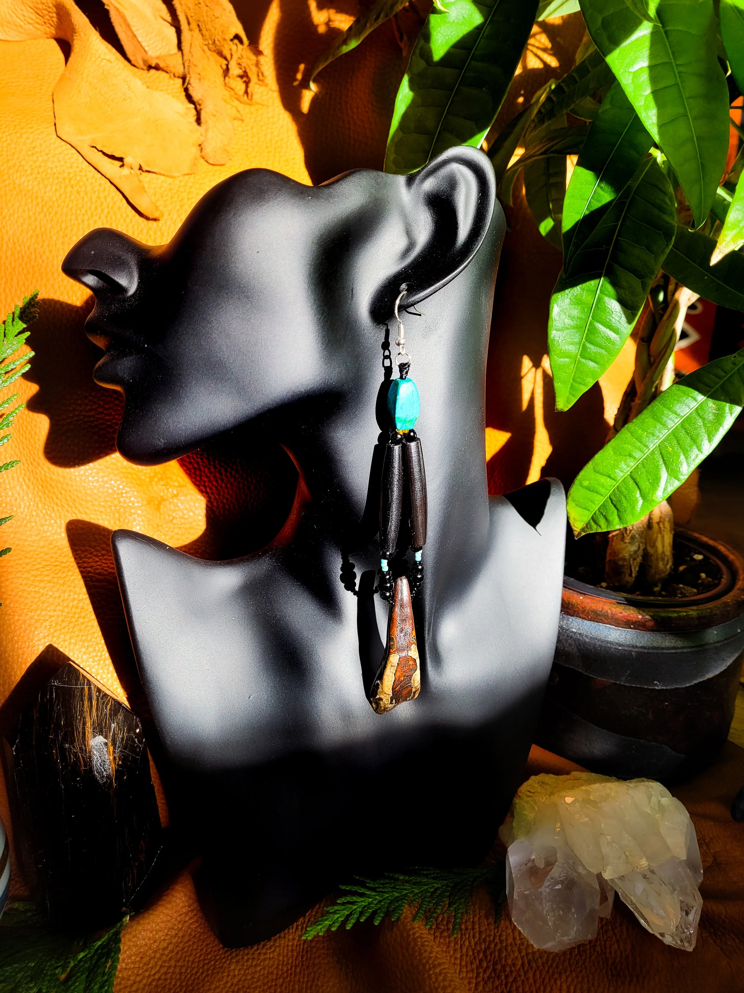Photo of long, heavy-weight Turquoise, Bone, and Buffalo Tooth earrings displayed on a dark jewelry mannequin with tan leather, crystals, pottery, and a green plant in the background. Earrings contain two dark brown bone pipe beads, Buffalo tooth, re