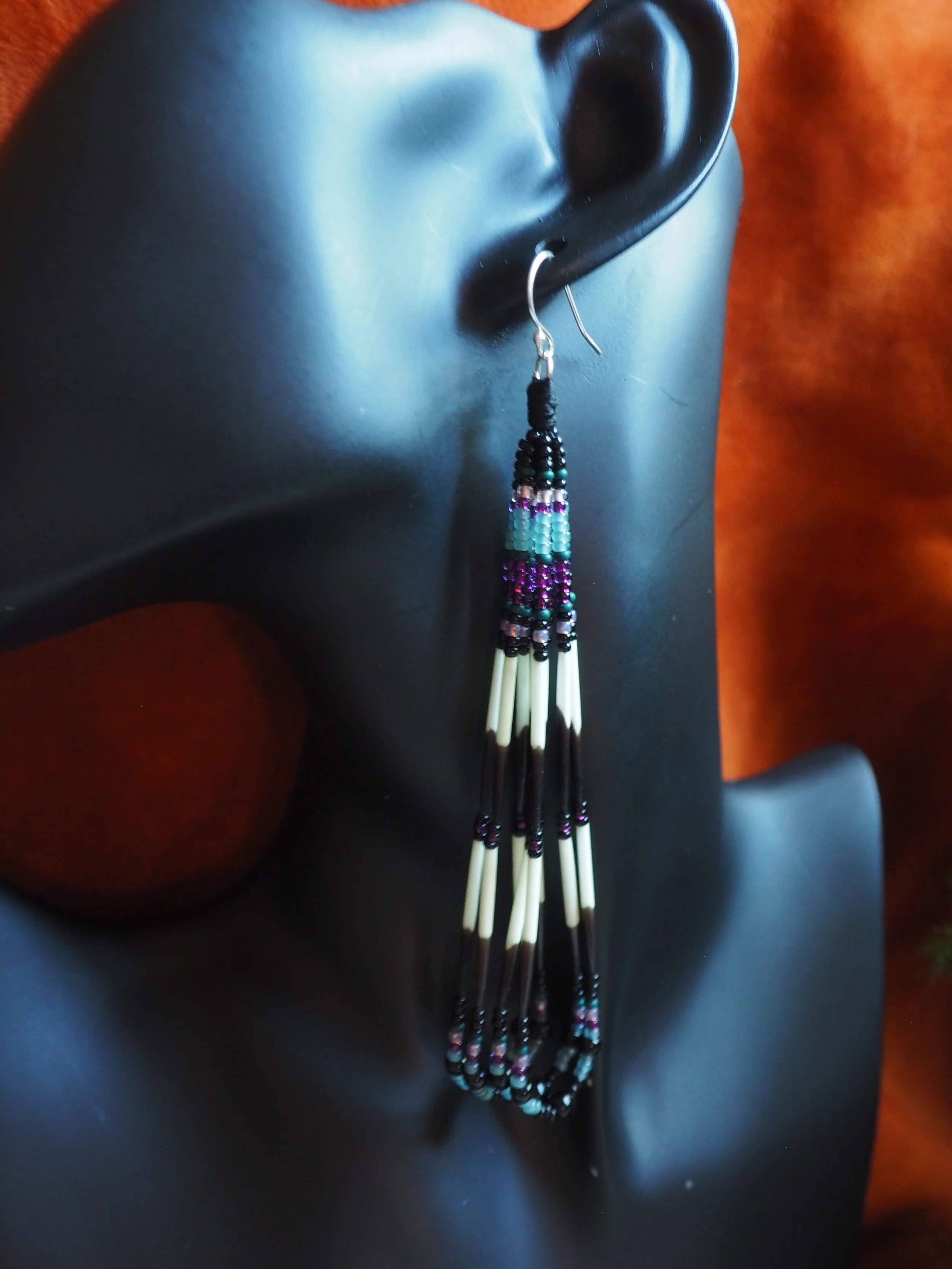 Photo of  long, lightweight traditional Ancestral Wabanaki Porcupine Quill earring hung on a dark jewelry mannequin in front of dark terra cotta colored leather backdrop. Earring contains 8 strands in four looped segments with two tiers of 16 white a