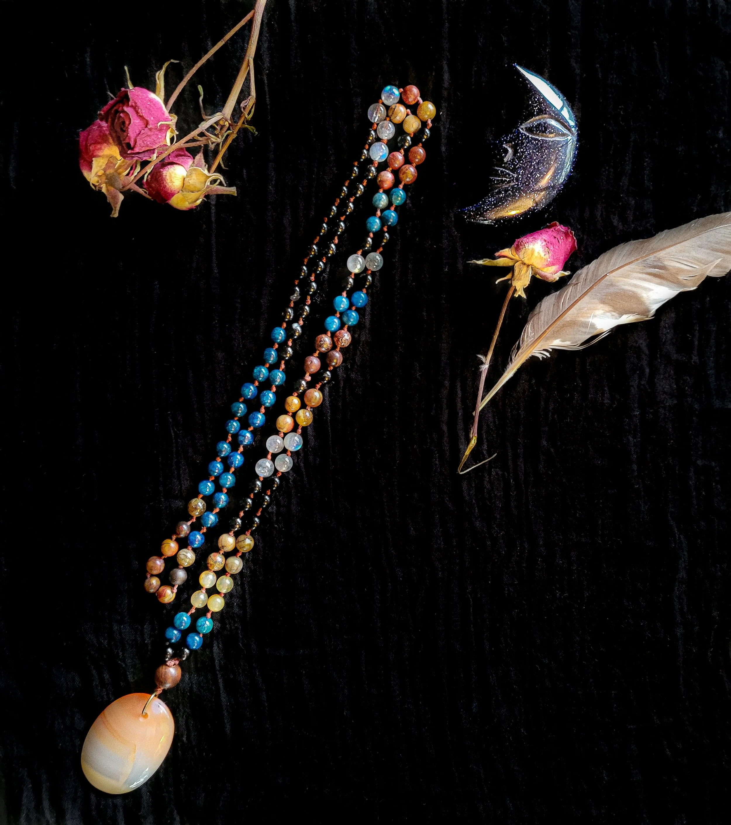 Photo of hand holding custom-made limited gemstone mala necklace with 8mm labradorite, lapis lazuli, crazy horse Jasper, tourmaline; 10mm sandalwood bead; and carnelian pendant. Feather, crystal, and dried roses are in the background. Commission desi