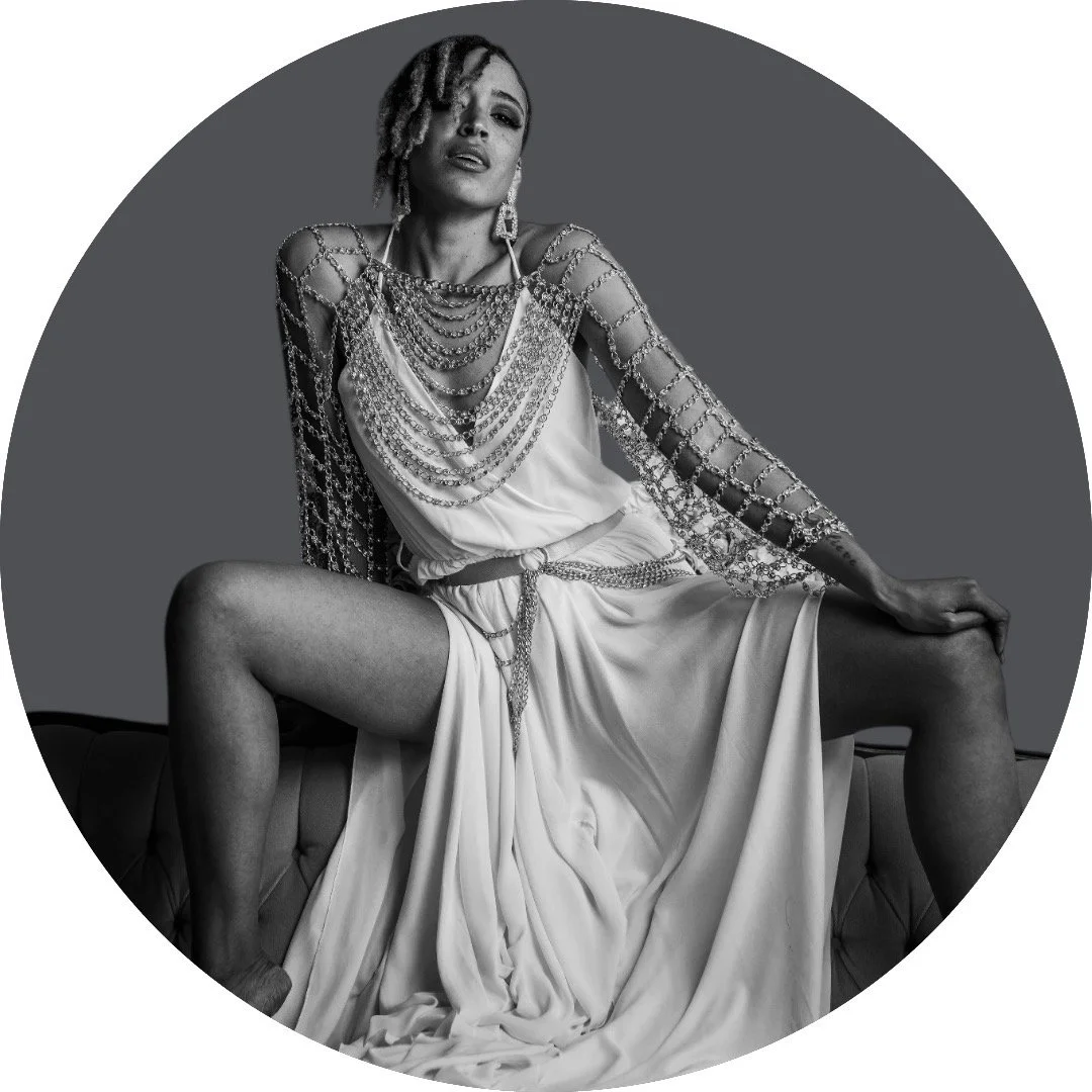 A woman sitting with her legs apart, wearing a flowing dress and multiple layers of jewelry, in a black and white photograph.