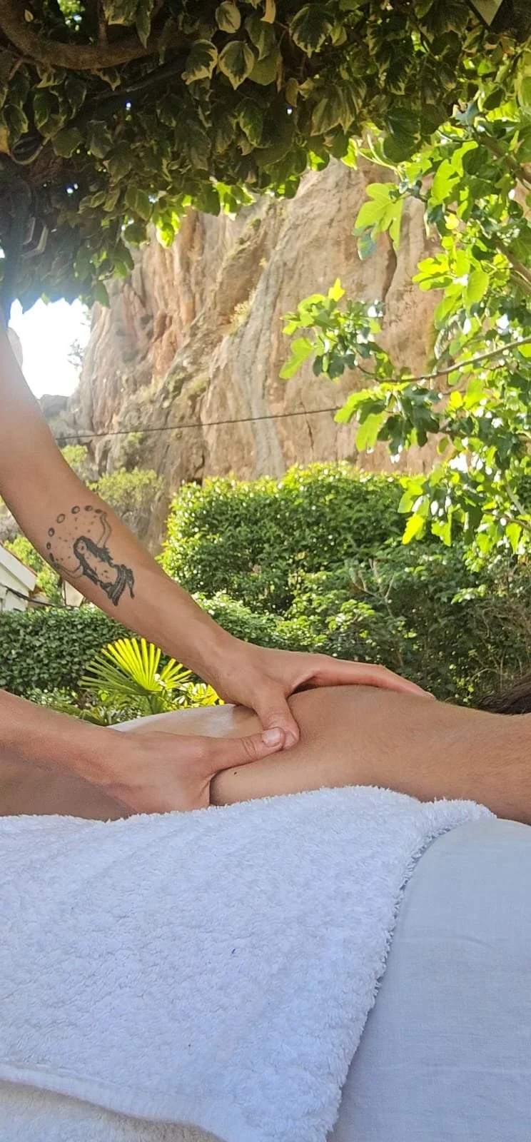 A person receiving a massage outdoors under a tree with green leaves, near a rocky cliff, with a tattoo of a mermaid on their arm.