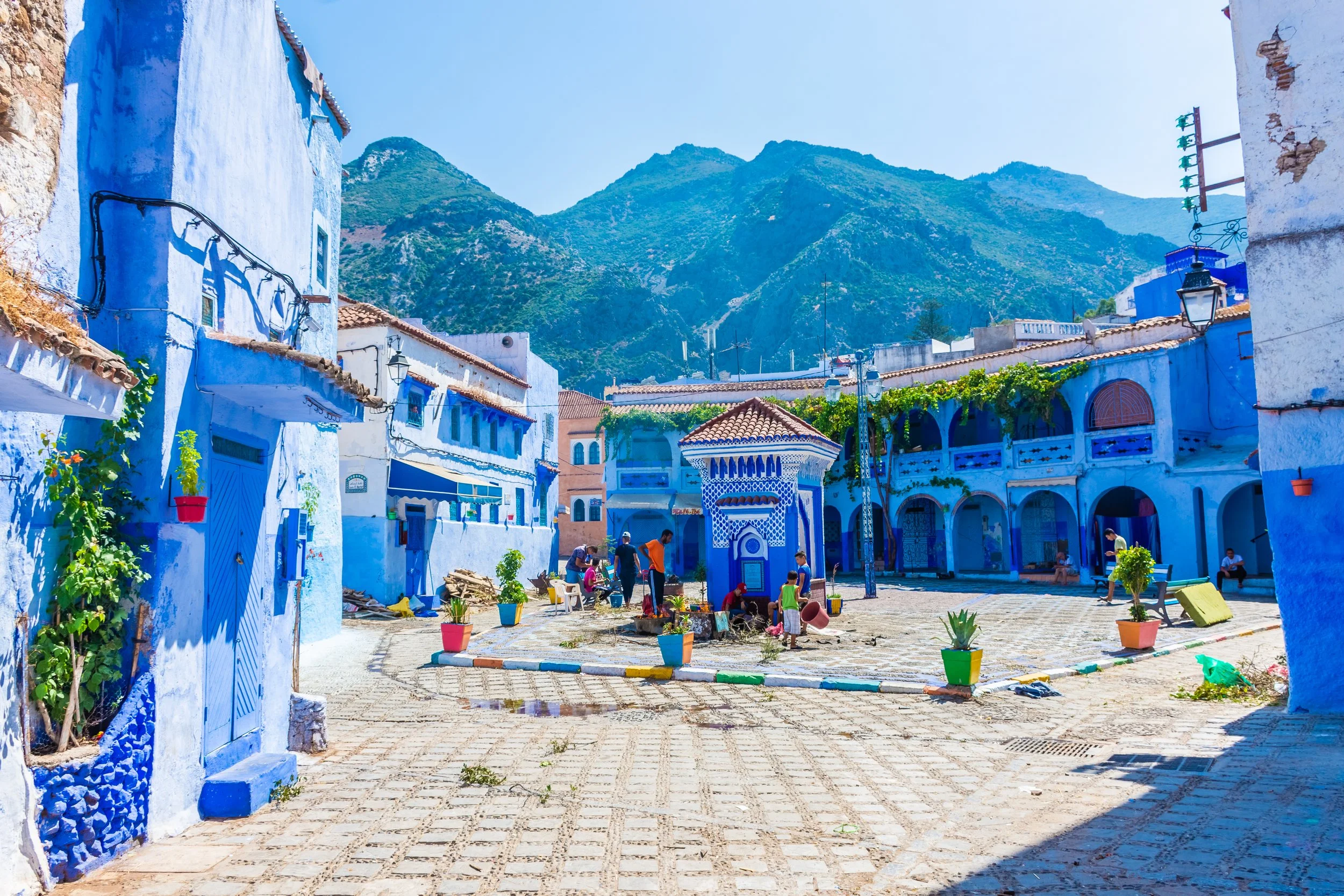 Blue-painted buildings surrounding a central plaza decorated with potted plants, with mountains in the background.