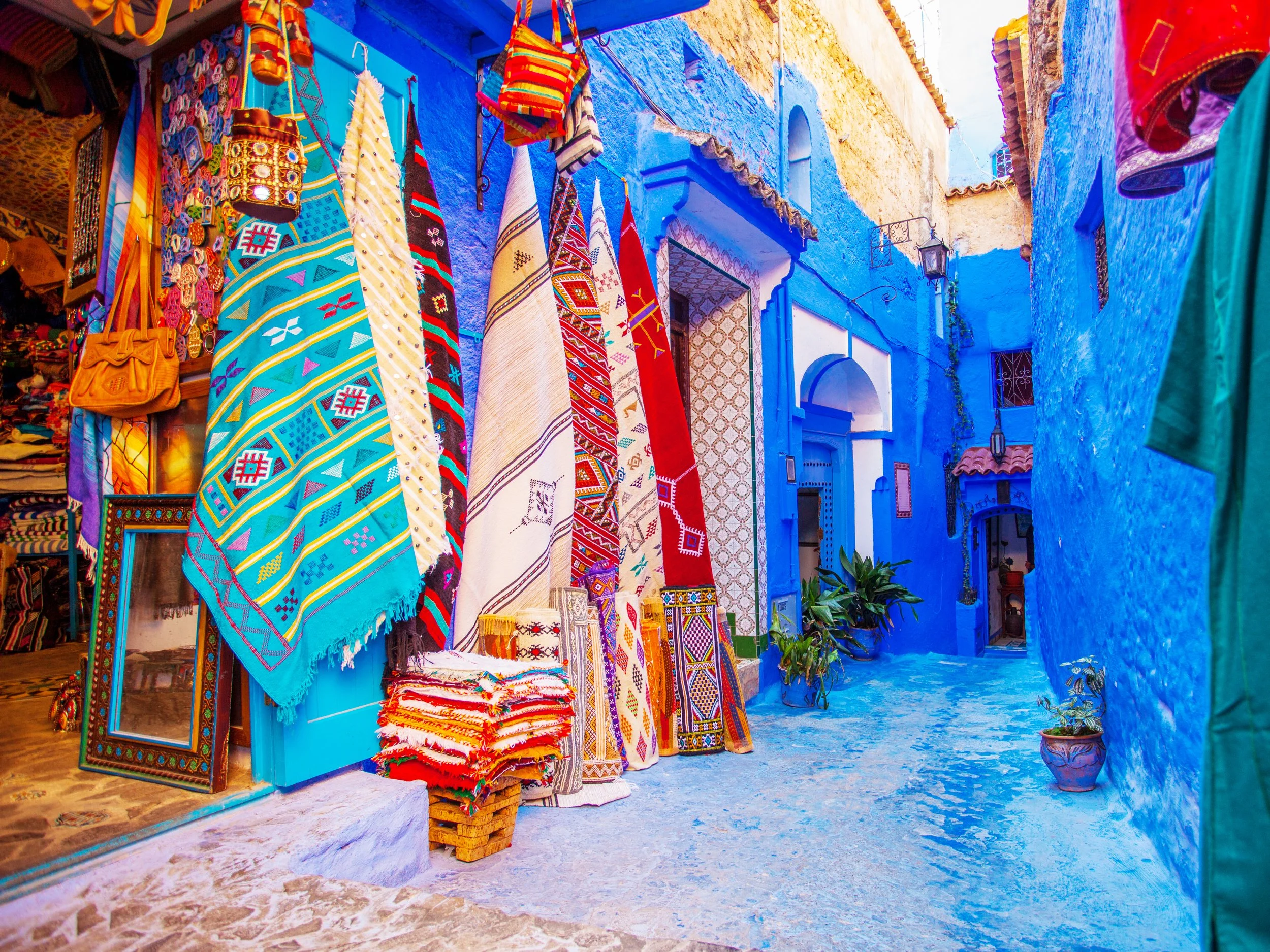 Colorful marketplace with hanging textiles and rugs, surrounded by vibrant blue walls and narrow street with potted plants.