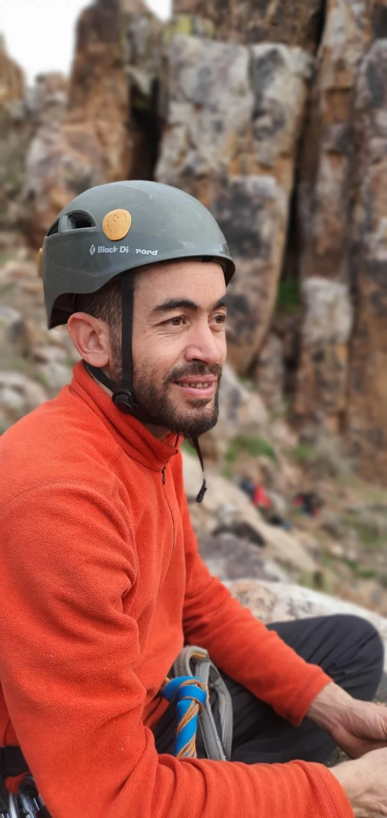 A man wearing a helmet and orange fleece jacket sitting on rocky terrain, ready to take beginner climbers to a multipitch climbing course.