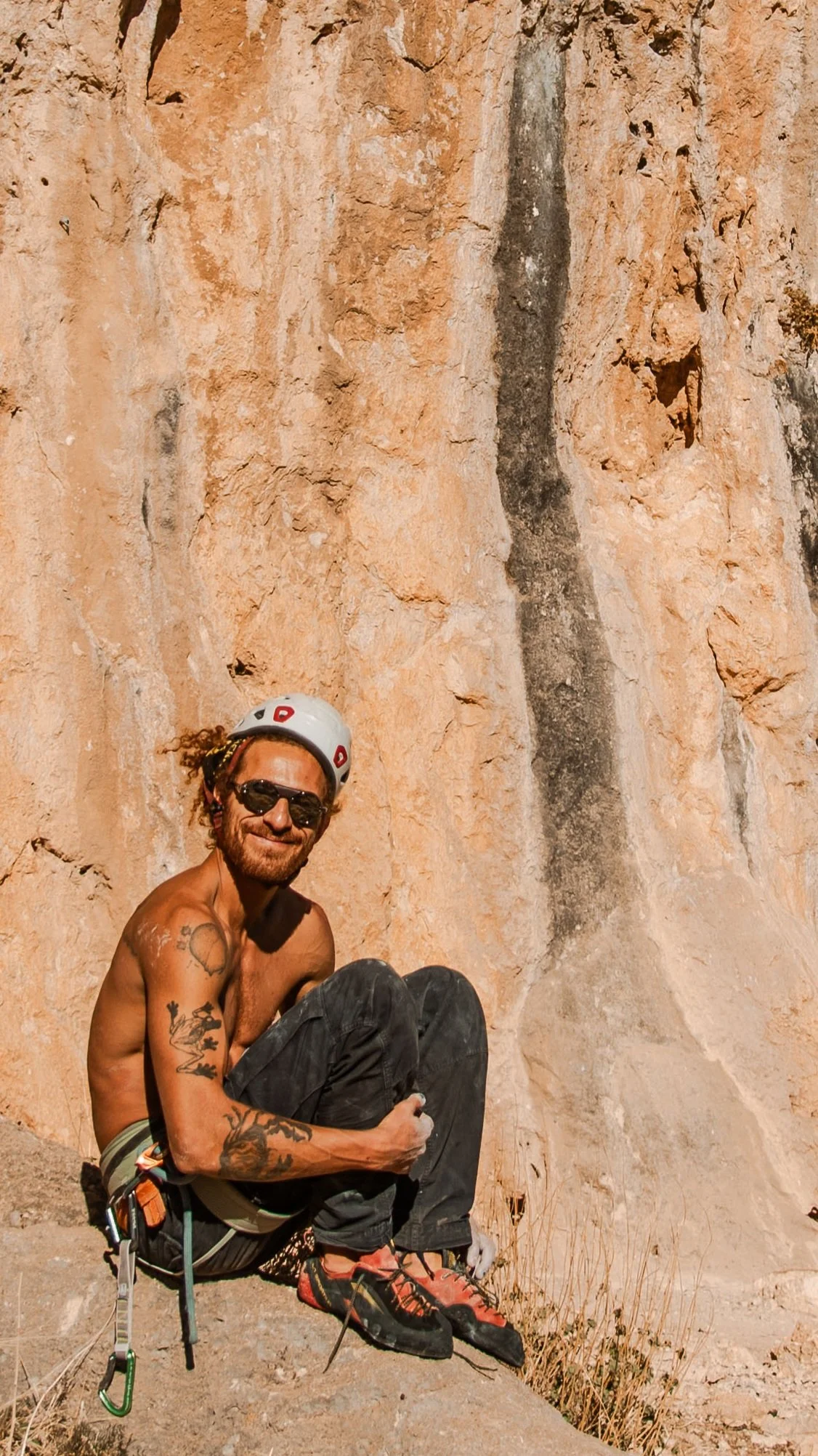 A man with tattoos, sunglasses, and a helmet sitting on a rock at the base of a rocky cliff, smiling at the camera. Enjoying the winter sun, perfect conditions for climbing in Morocco.