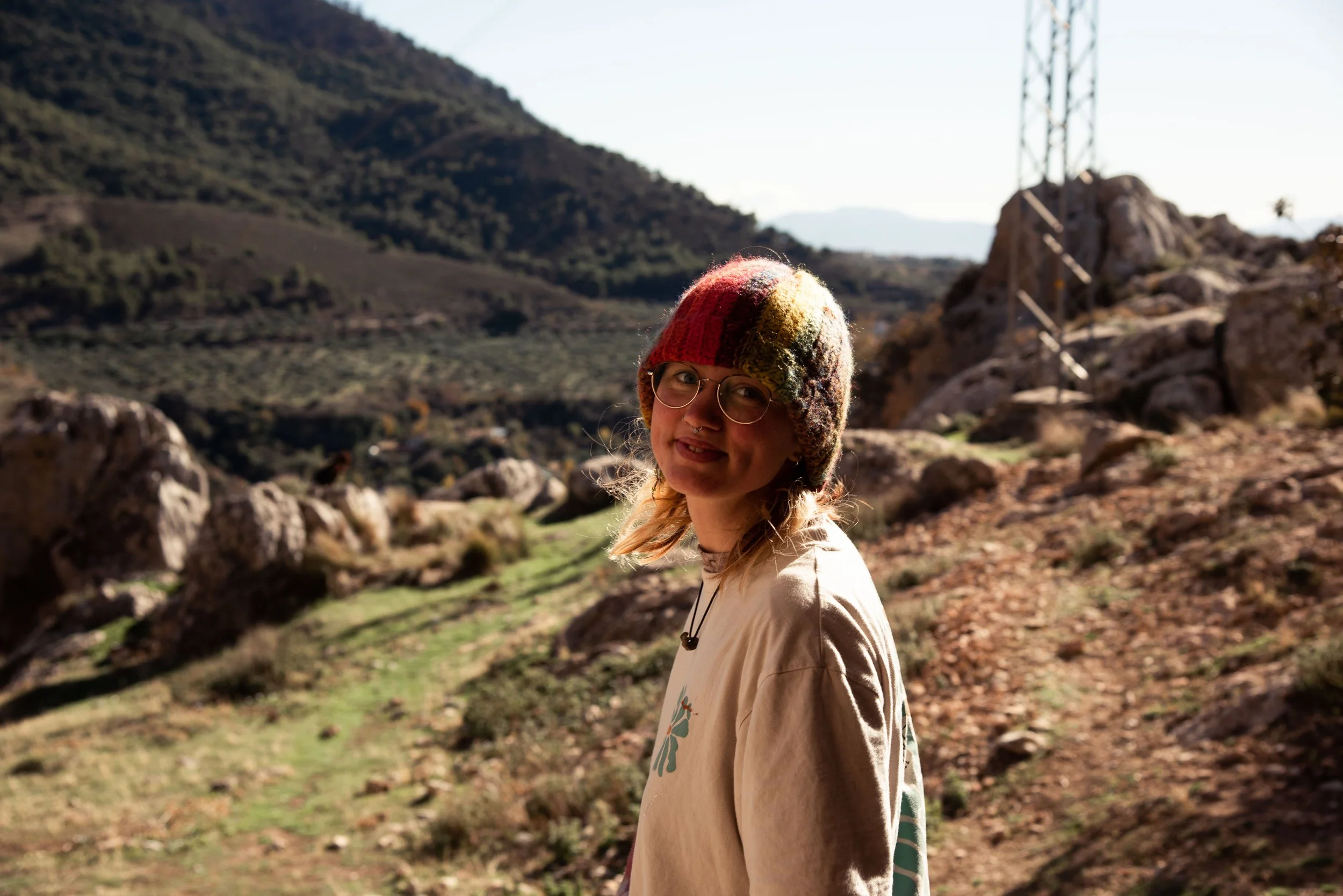 Young woman wearing glasses and a colorful knit hat stands outdoors on a trail in a mountainous landscape with rocks and green vegetation, with hills and a tower in the background.