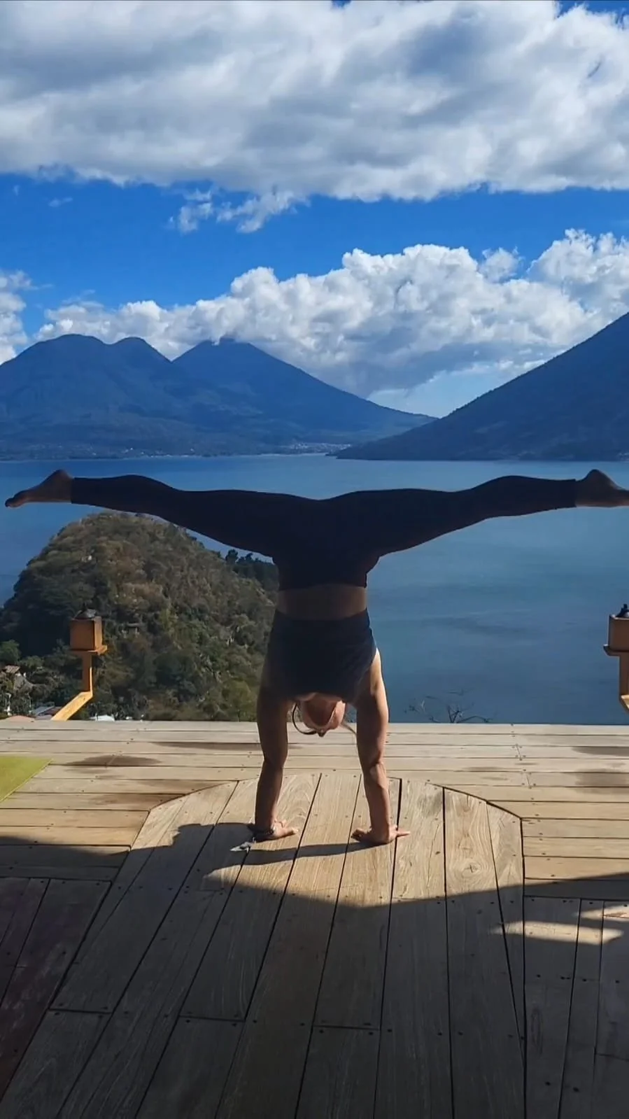 A person doing a handstand on a wooden deck with a scenic view of mountains, lake, and a partly cloudy sky in the background.