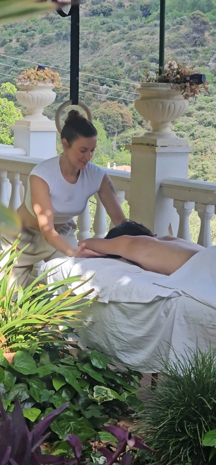A woman giving a massage outdoors on a covered patio with a scenic hillside view, large decorative planters, and lush green plants in the foreground.