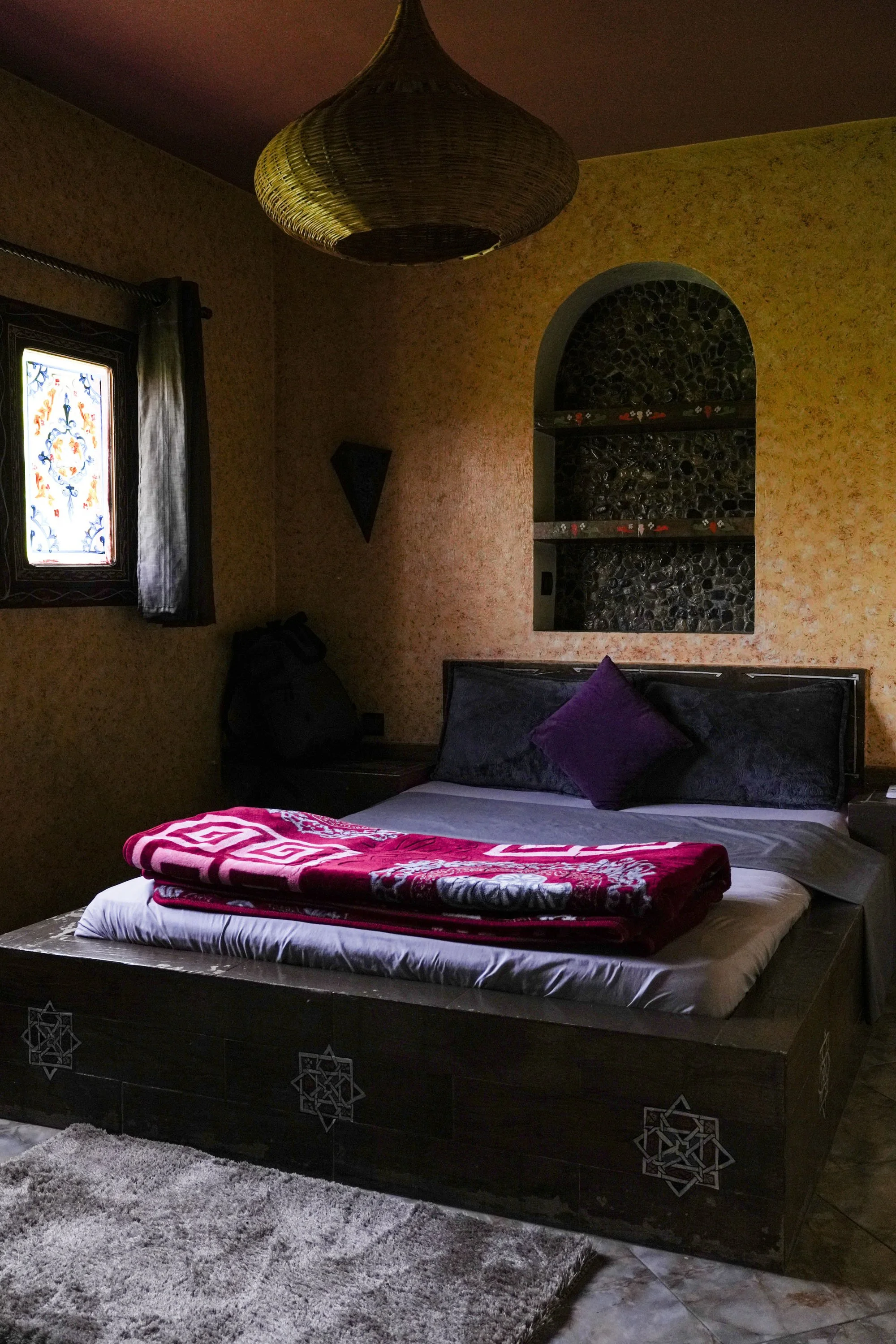 A bed in a dimly lit bedroom with a purple pillow and a red blanket, dark wall decor, and a small window with stained glass.