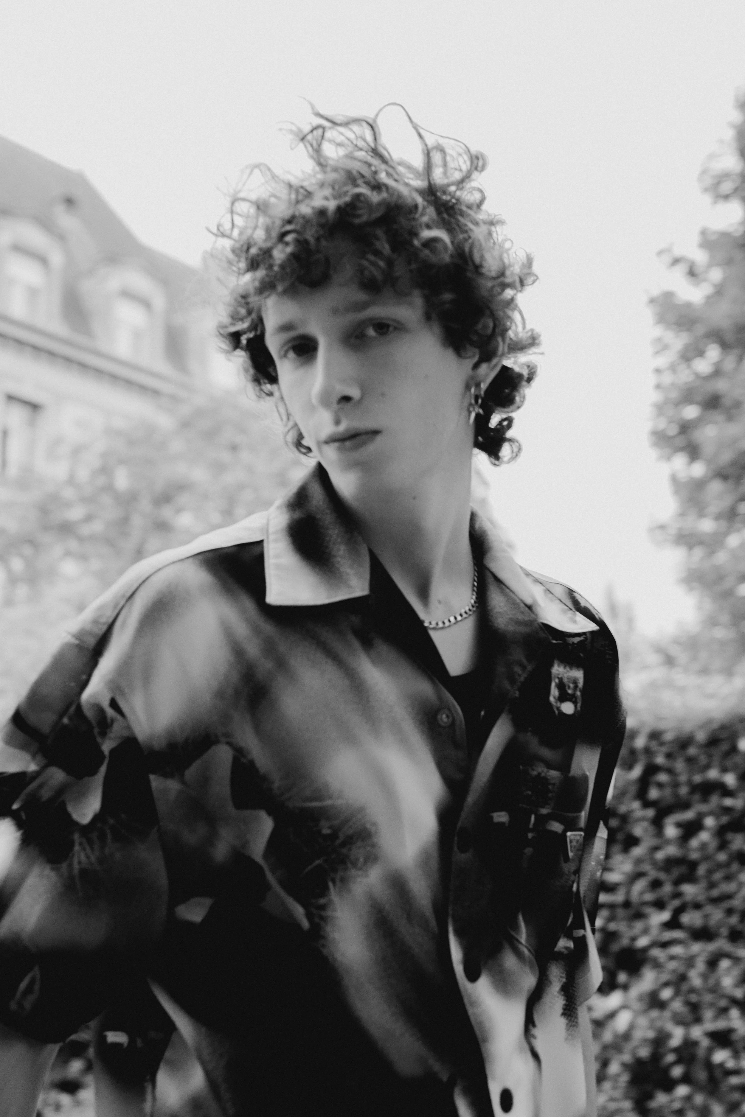Jeune personne avec des cheveux bouclés et courts, portant une veste à motifs et une chaîne, posant à l'extérieur, arrière-plan flou de bâtiments et d'arbres.