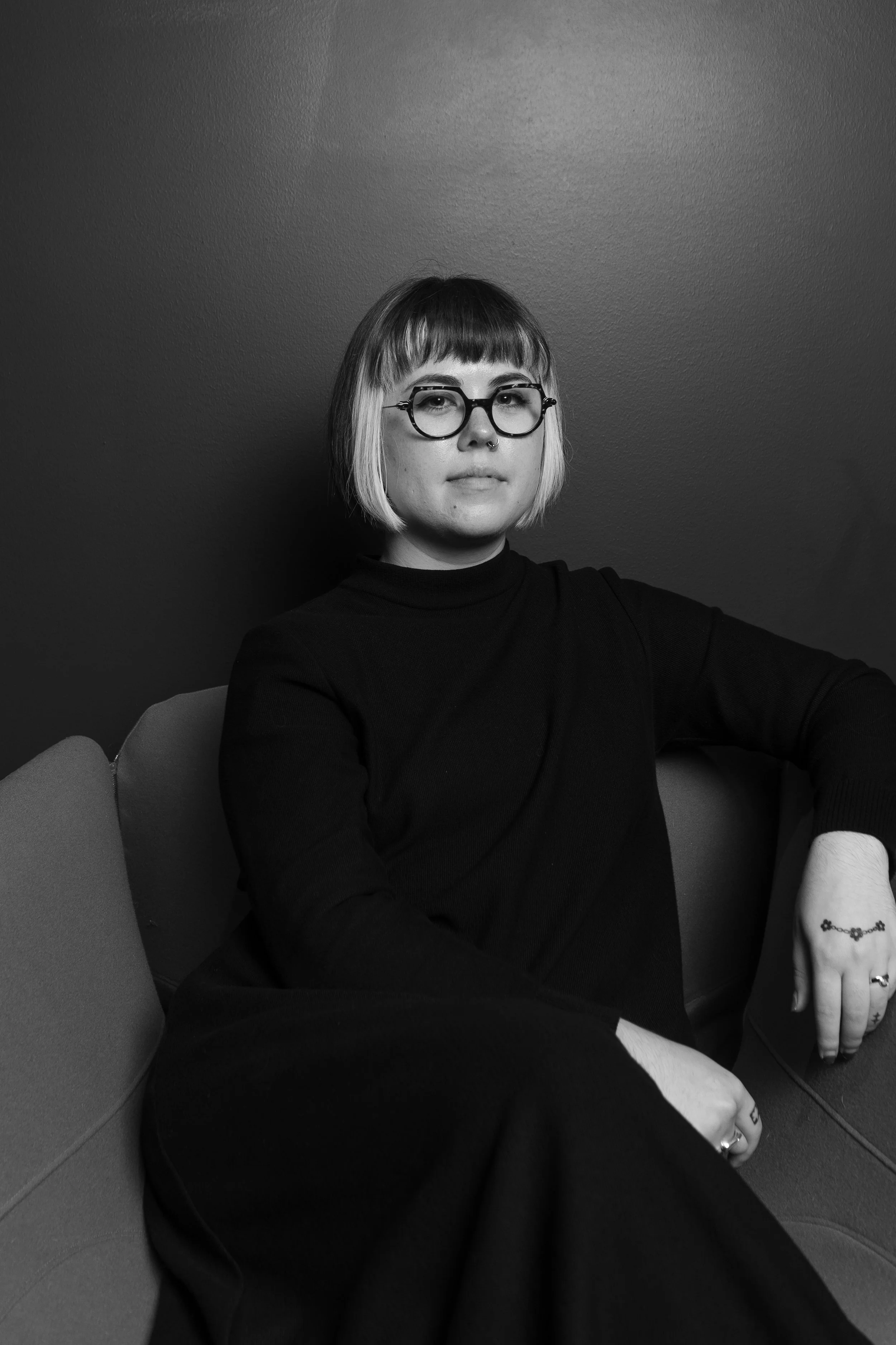 A young woman with short bob hairstyle, wearing glasses and a black turtleneck, sitting on a couch against a dark wall, with tattoos on her fingers and hand in a black-and-white photo.