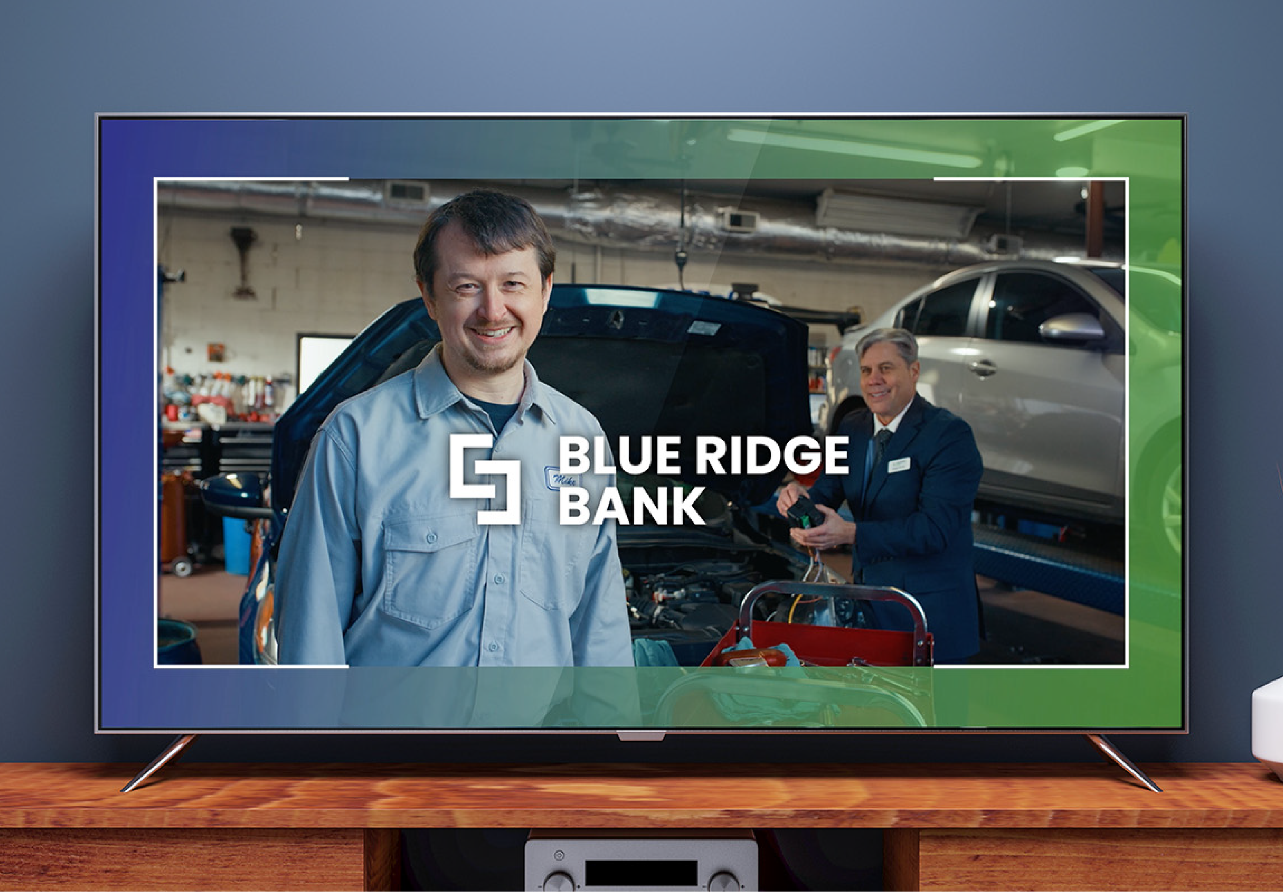 Television screen displaying a Blue Ridge Bank advertisement with two mechanic or service technicians in an automotive workshop, one smiling at the camera and the other working on a vehicle.