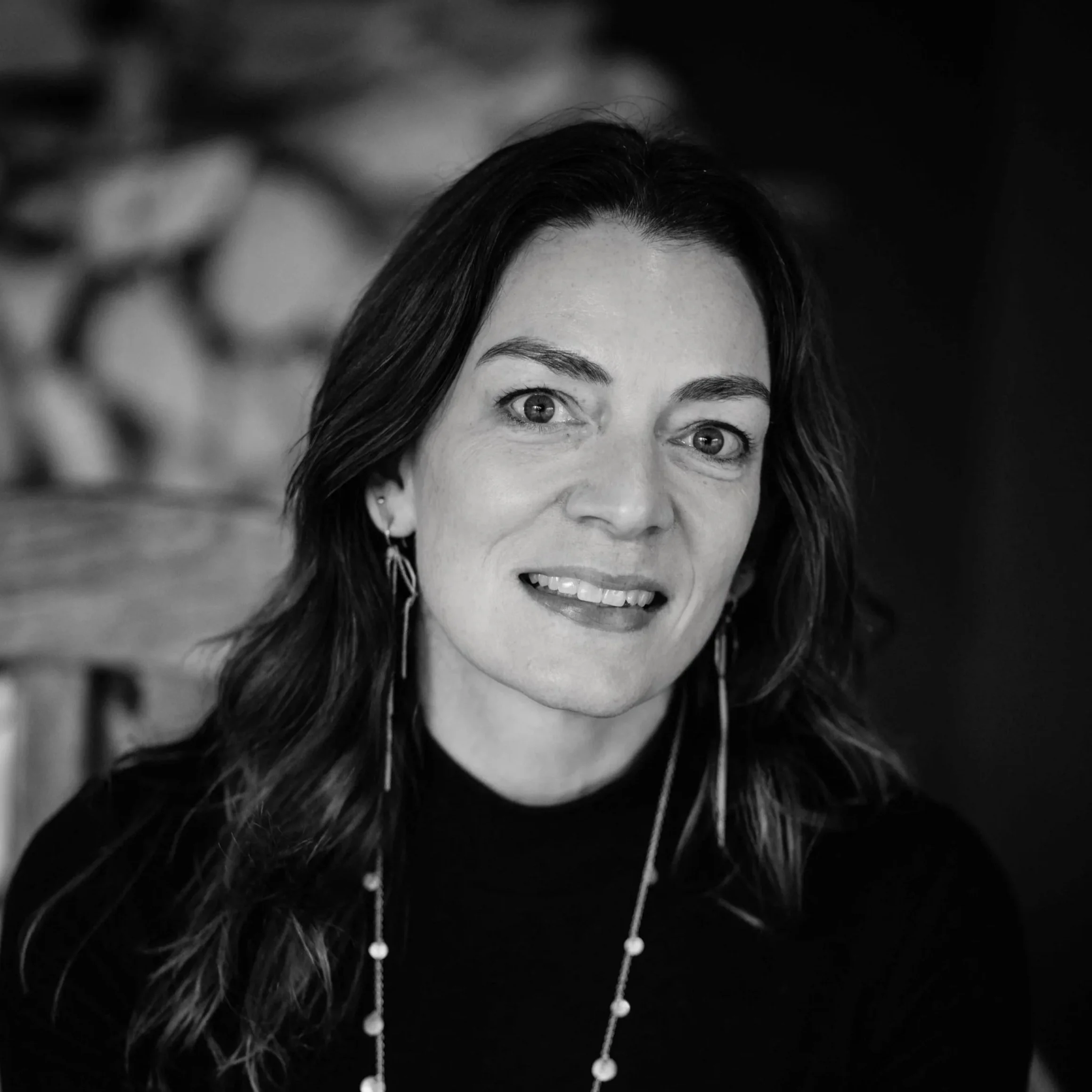 Black and white portrait of a woman with long dark hair, smiling slightly, wearing earrings and a necklace.