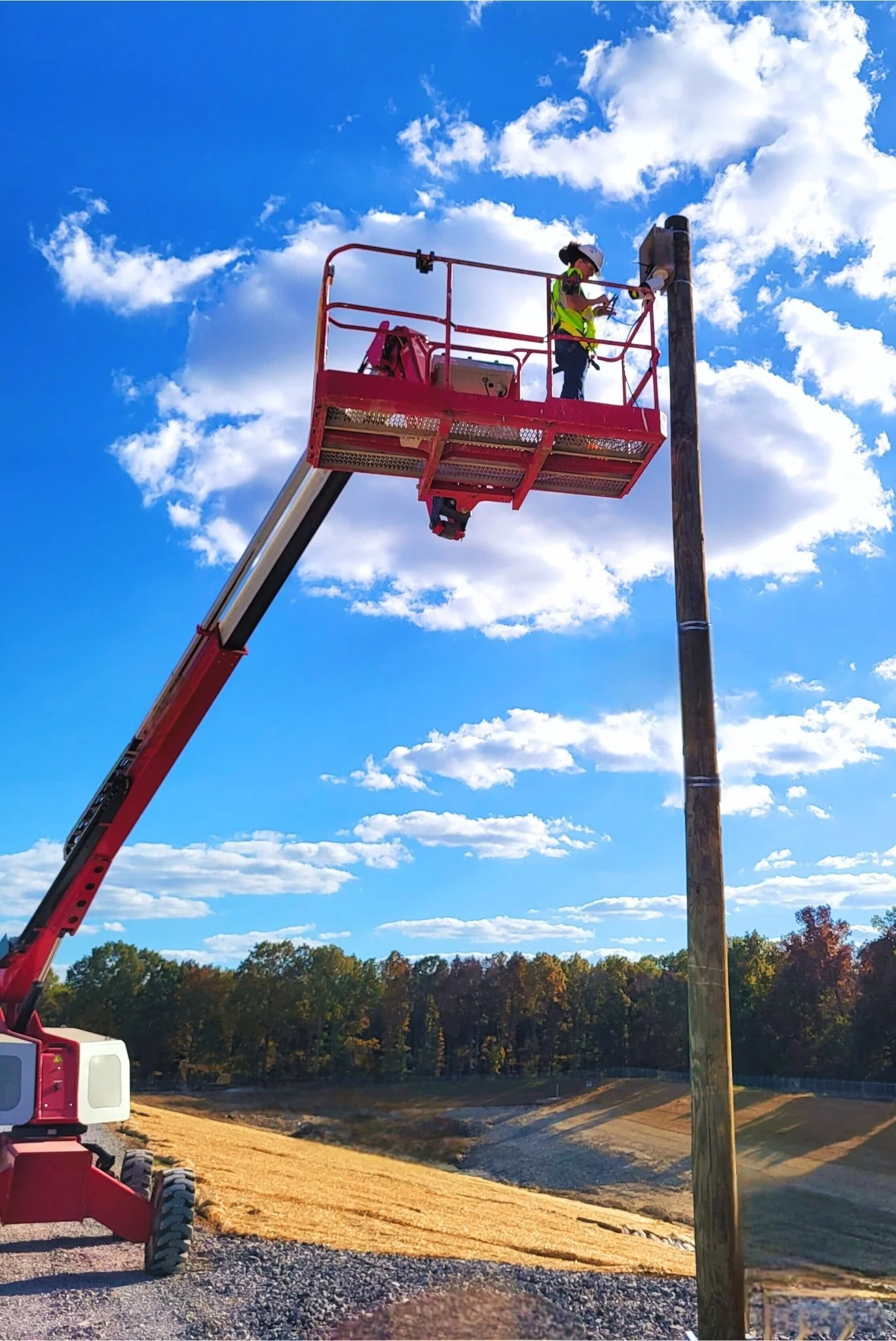 worker in hard hat and vest stands in basket of red boom lift to service security cameras on a wooden telephone pole