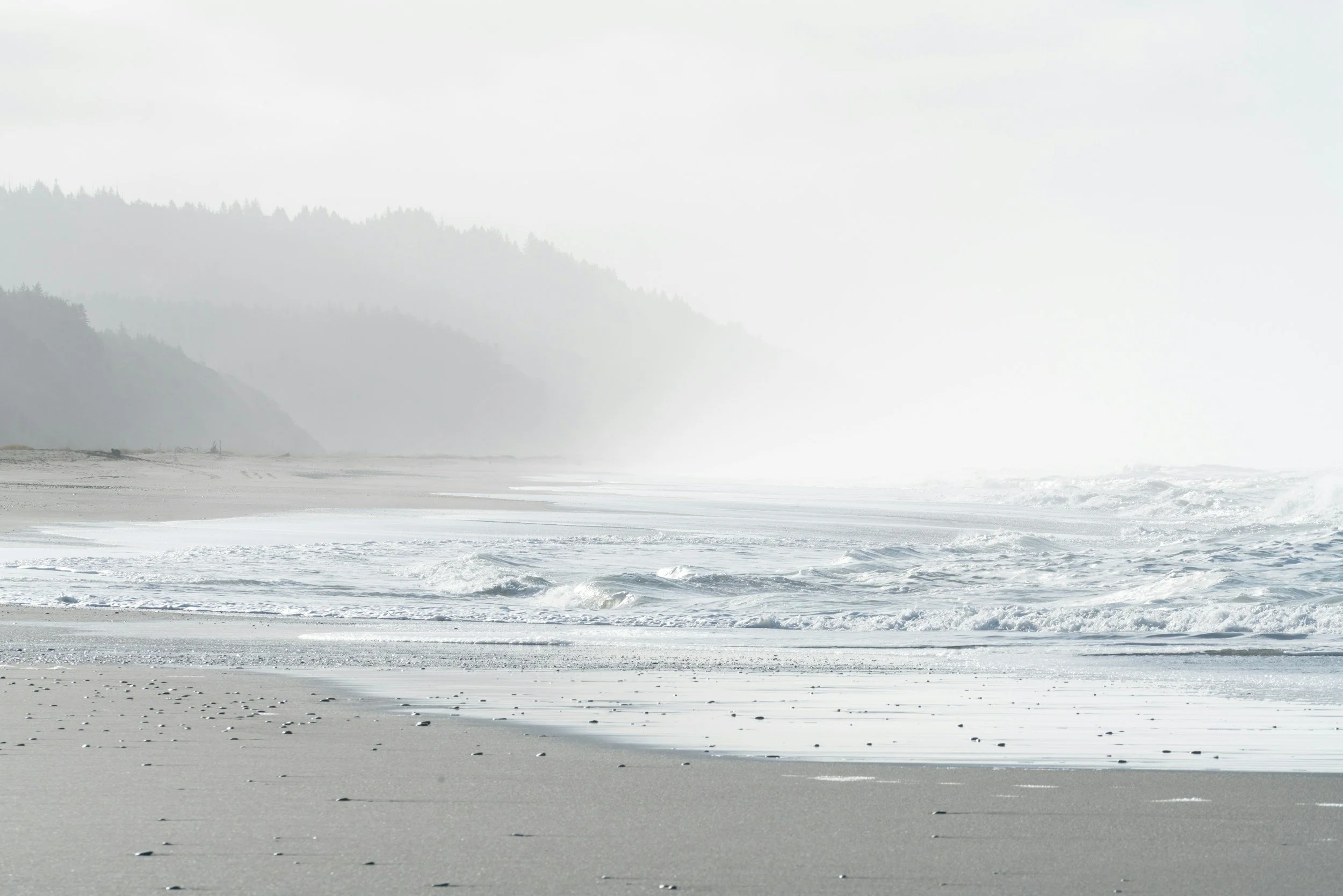 Bright, foggy beach with gentle waves and distant forested hills