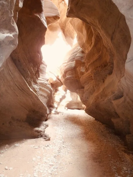 Narrow slot canyon with smooth, layered sandstone walls and a sandy floor, illuminated by sunlight from above.