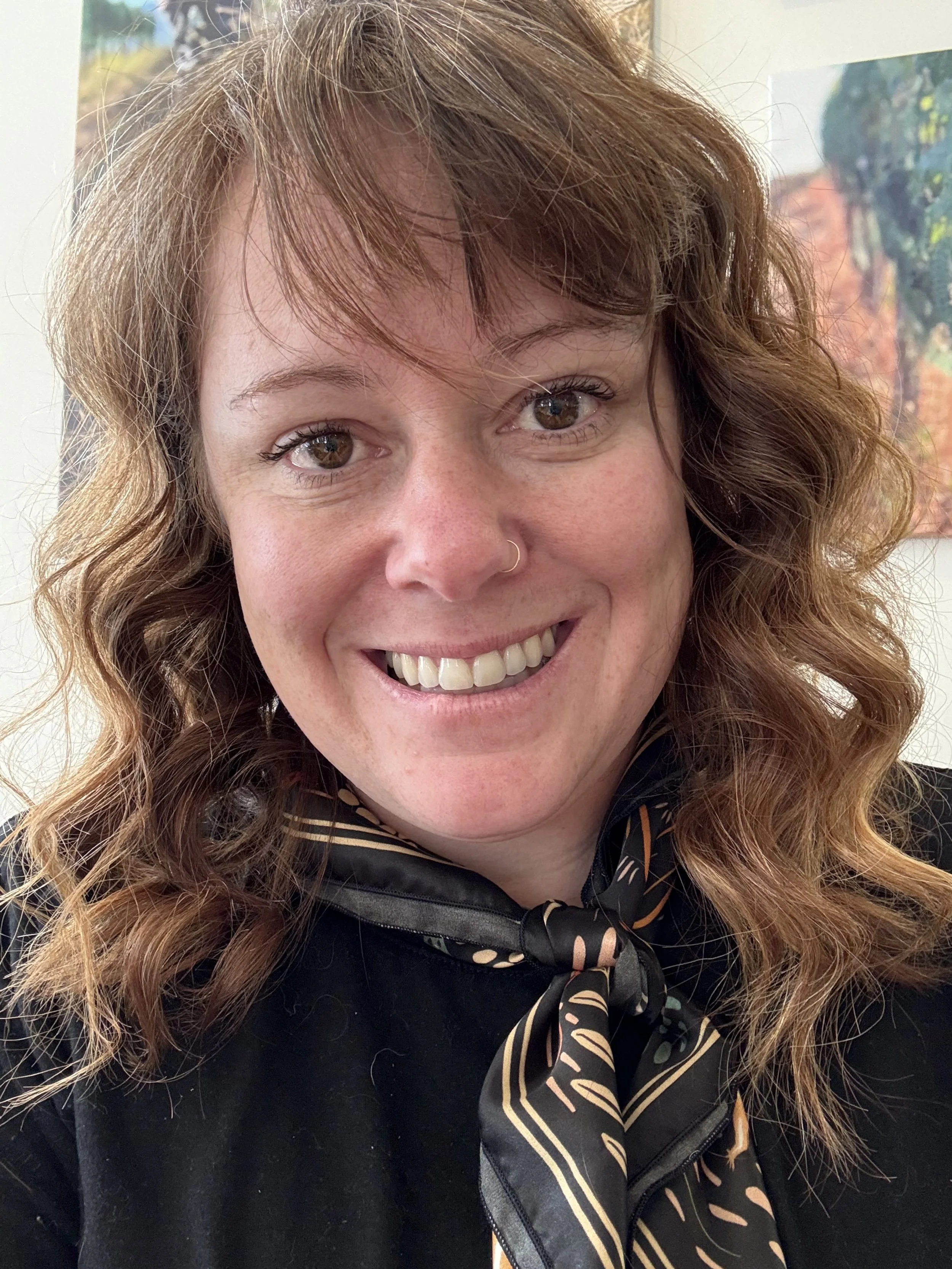 A woman with curly auburn hair smiling, wearing a black top and a patterned scarf, in front of colorful artwork.