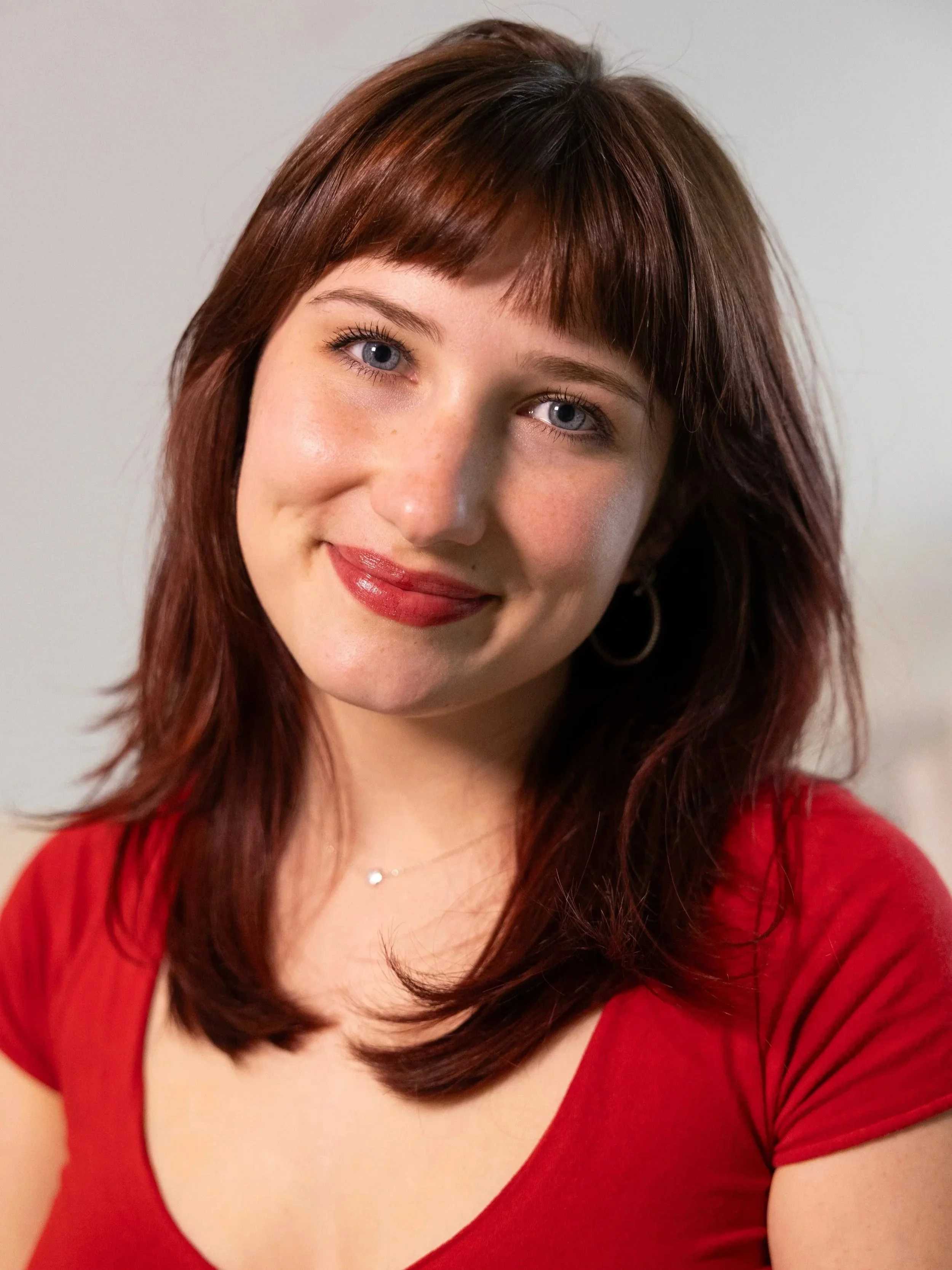 A woman with red hair, blue eyes, and light skin wearing a red top, standing against a plain white background.