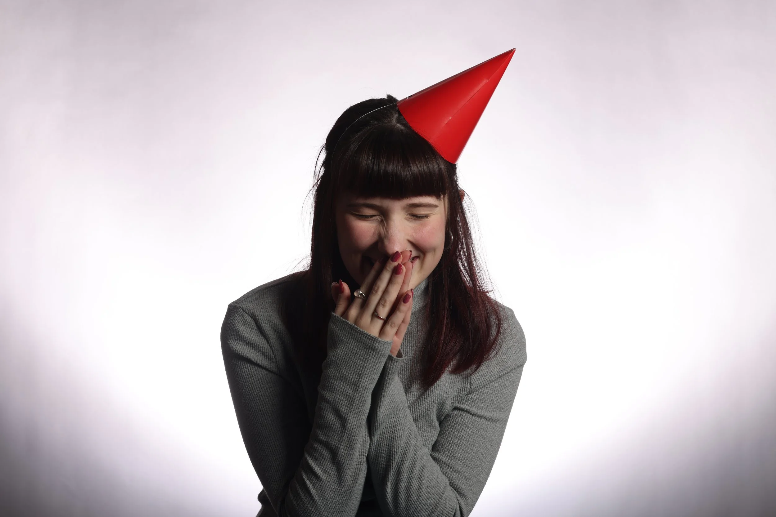A young woman laughing with her eyes closed, wearing a gray top and a red party hat, with her hand covering her mouth, against a plain white background.