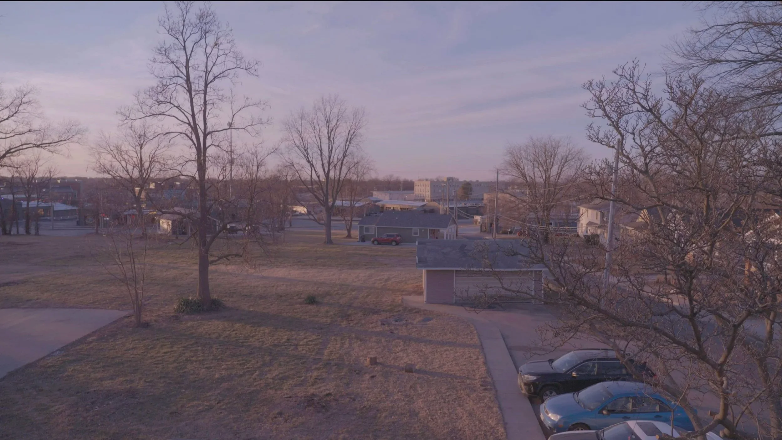 A suburban neighborhood park during late fall or winter with leafless trees, a grassy area, parked cars, and buildings in the background under a partly cloudy sky.