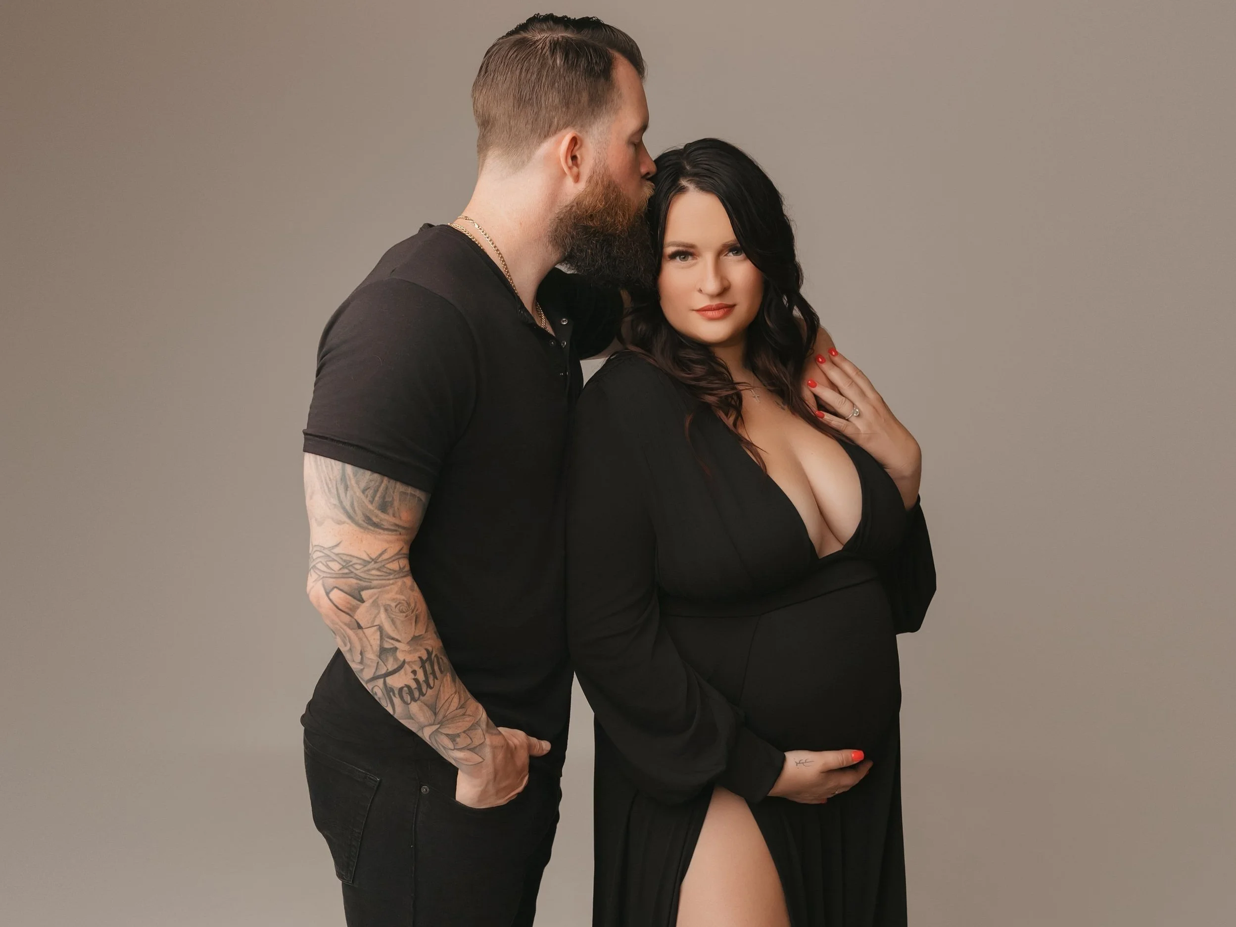 A man with tattoos and a beard is kissing a woman with black hair, who is wearing a black dress, against a plain gray background.