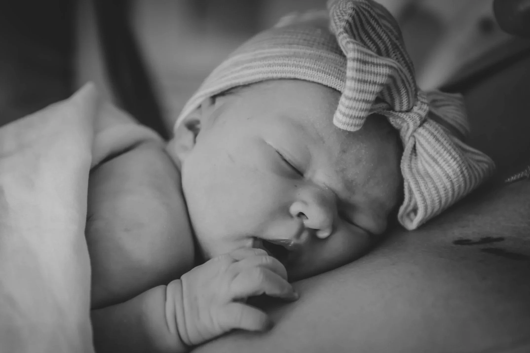 A sleeping baby with a striped hat, resting on a person's chest, with an adult's hand gently touching the baby's hand.