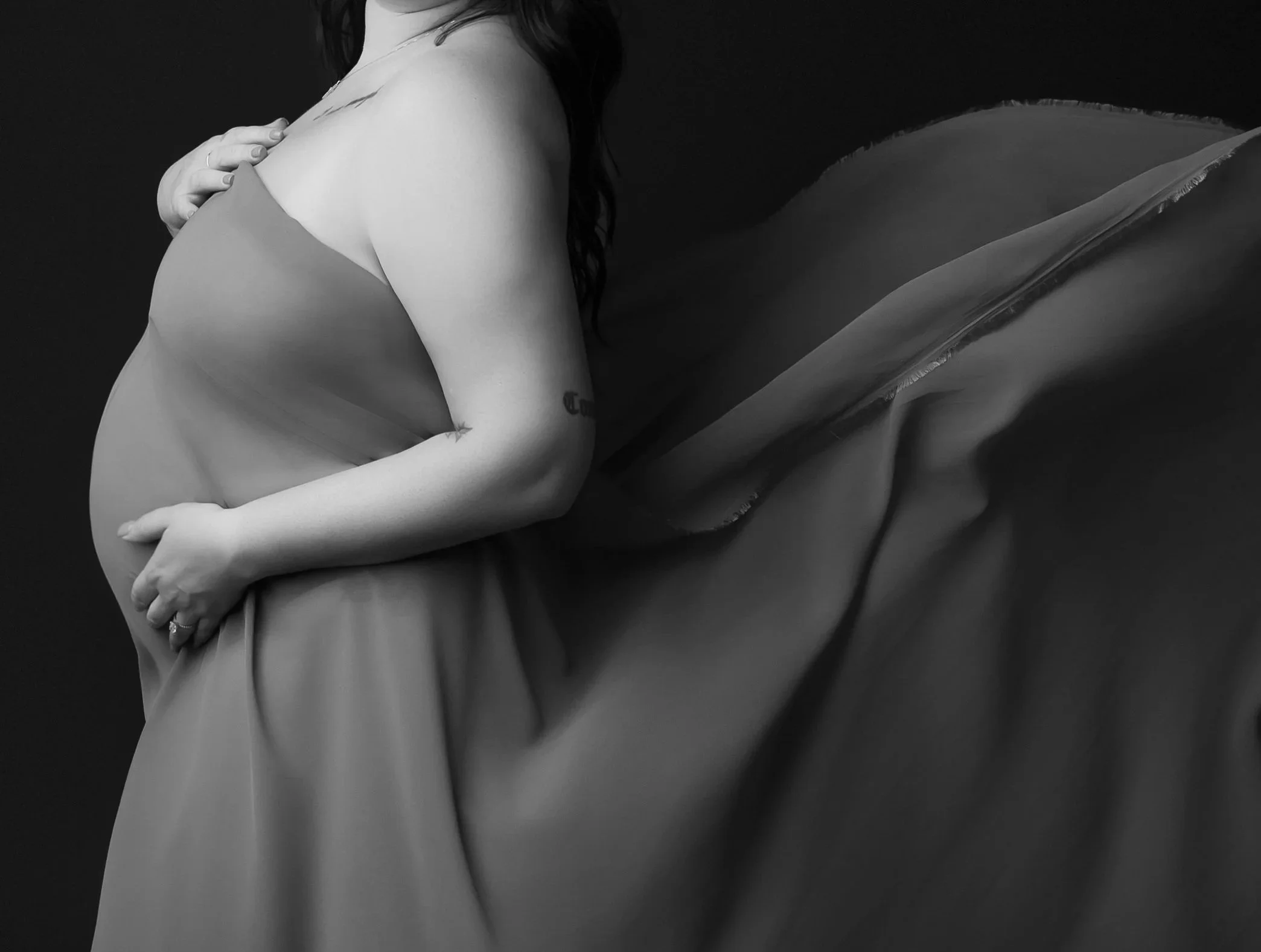A black and white photo showing a woman with long, wavy hair, sitting on a fabric surface. She is covering her chest with her arms, and is wearing a ring on her finger. The image captures her from the shoulders down, with a focus on the profile of her upper body.