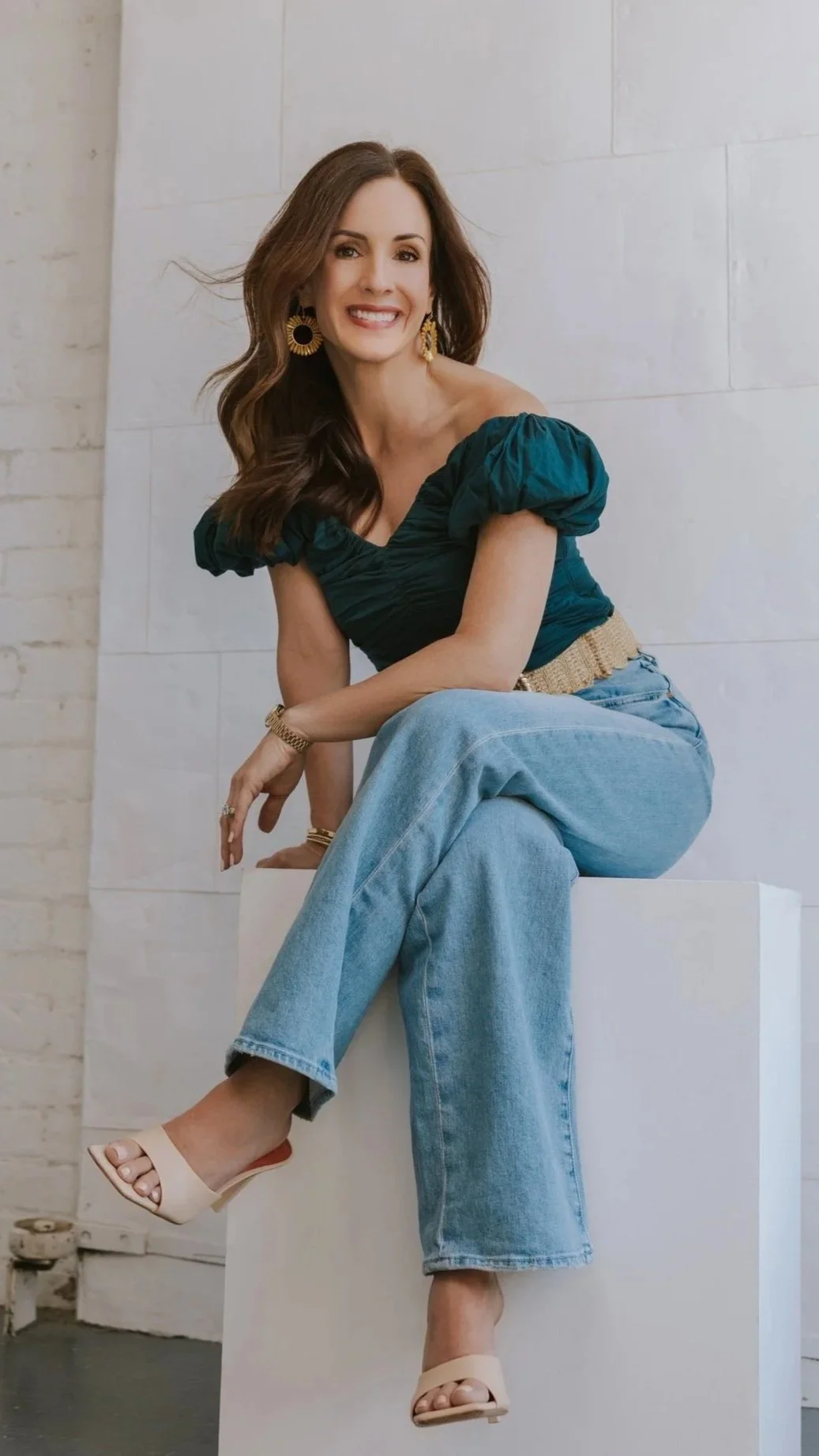 A woman with long dark hair, wearing a dark green top with puffed sleeves, light blue jeans, beige high heel sandals, and gold jewelry, is sitting on a white block and smiling at the camera in a minimalist indoor setting. Dr. Jennifer Uggen.