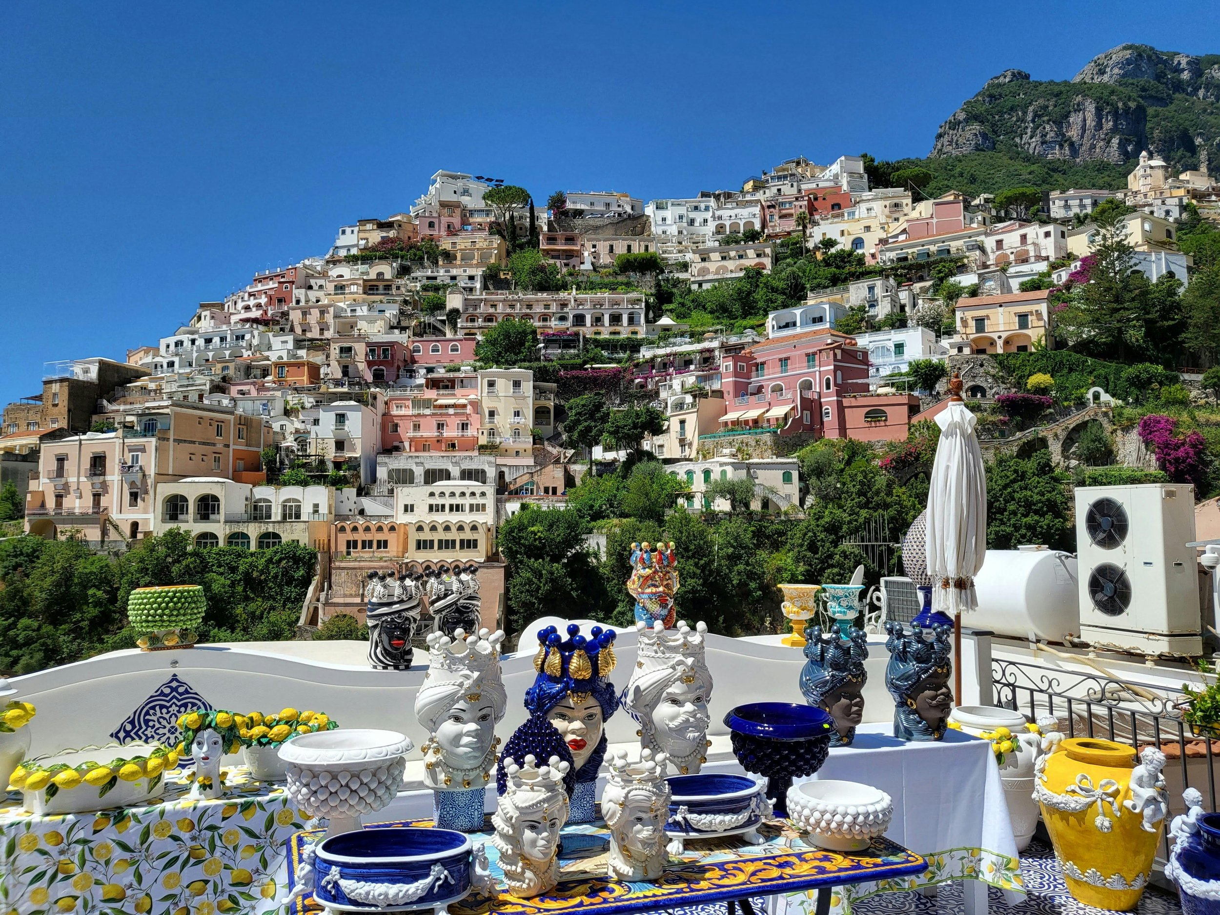 Colorful ceramics and decorative artifacts displayed on a terrace with a hillside town of pastel-colored houses in the background and a clear blue sky.