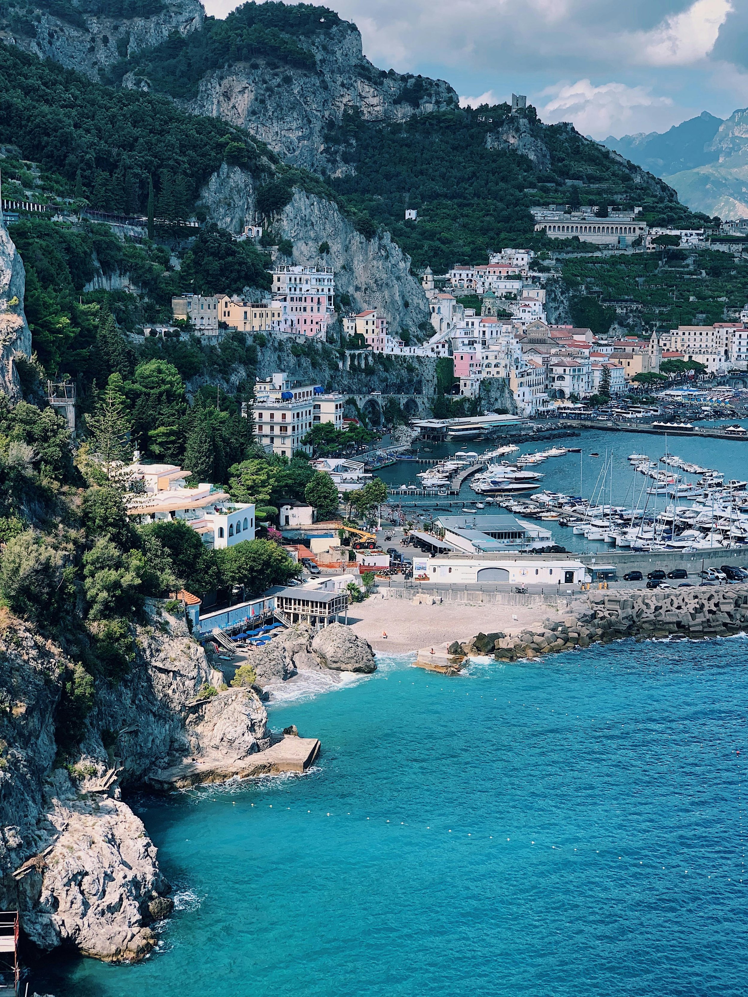 Coastal hillside town with colorful buildings and a marina filled with boats, overlooking turquoise water with rocky shoreline.