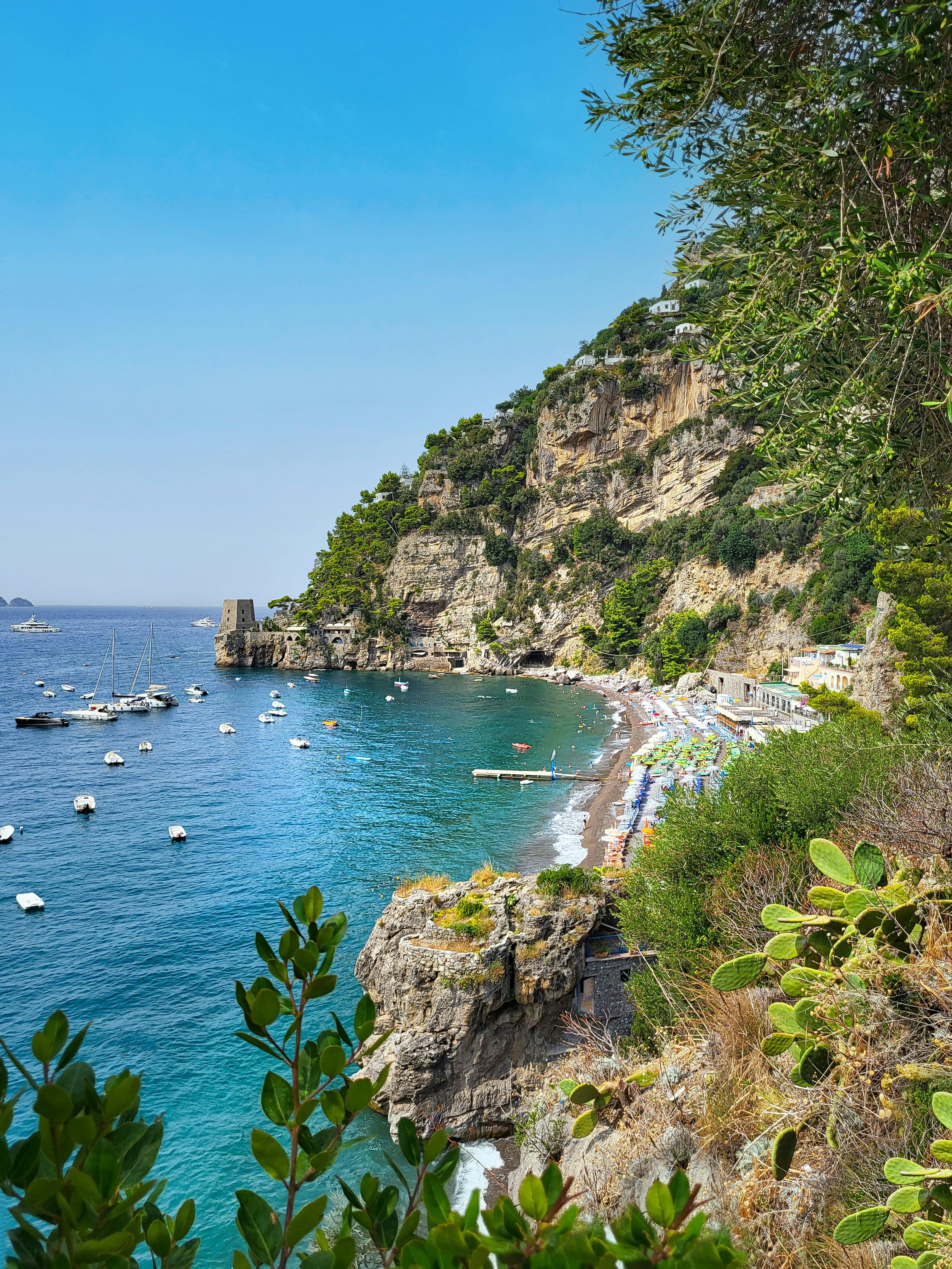 A scenic coastal scene with a rocky hillside, lush green vegetation, a small beach with colorful umbrellas, and boats anchored in the turquoise water.