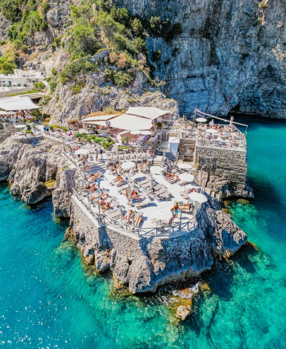 A rocky seaside restaurant and bar built on a cliff with turquoise water below, umbrellas, sunbathers, and people enjoying a sunny day.