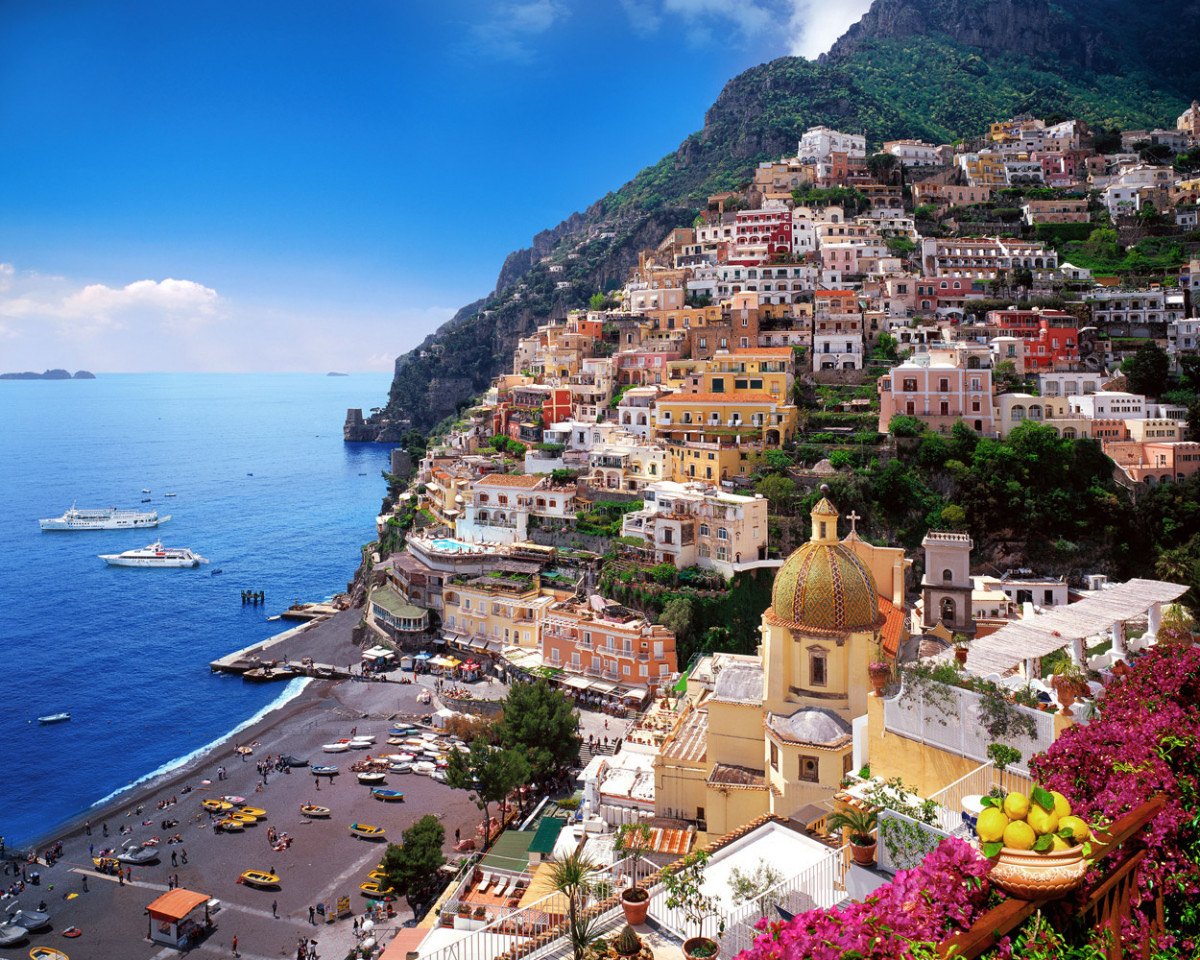 Colorful hillside village with tightly packed buildings, a church with a domed roof, a beachfront with boats, and the sea in the background.