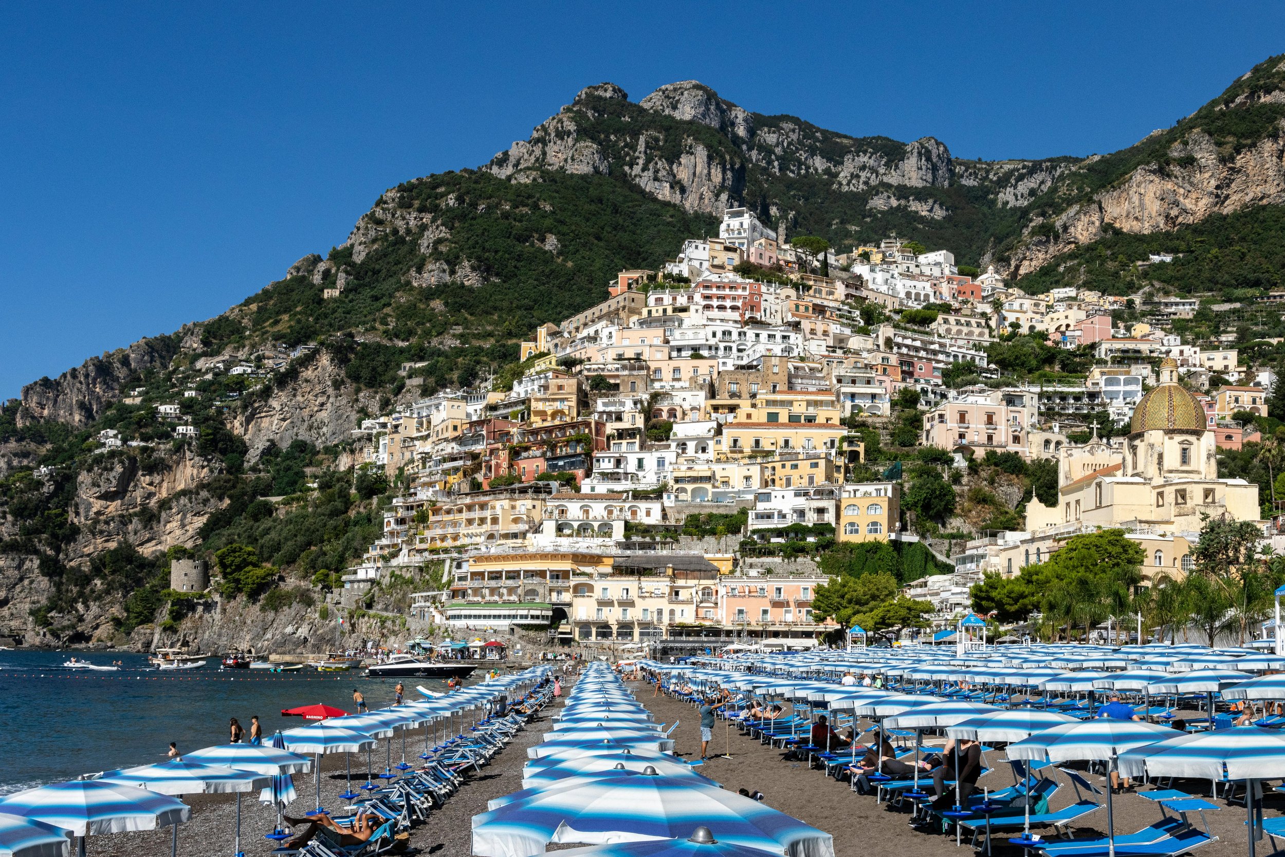 A beach with rows of blue and white umbrellas and lounge chairs, with colorful buildings on a hillside and mountains in the background under a clear blue sky.