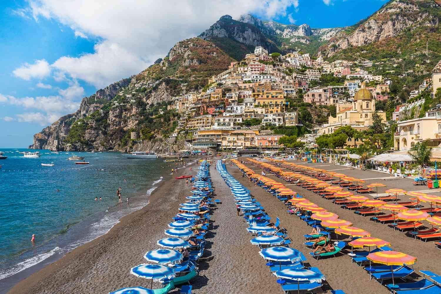 Colorful beach with rows of blue and orange umbrellas and lounge chairs along a rocky shoreline, with boats in the water and colorful hillside buildings in the background in Amalfi Coast, Italy.